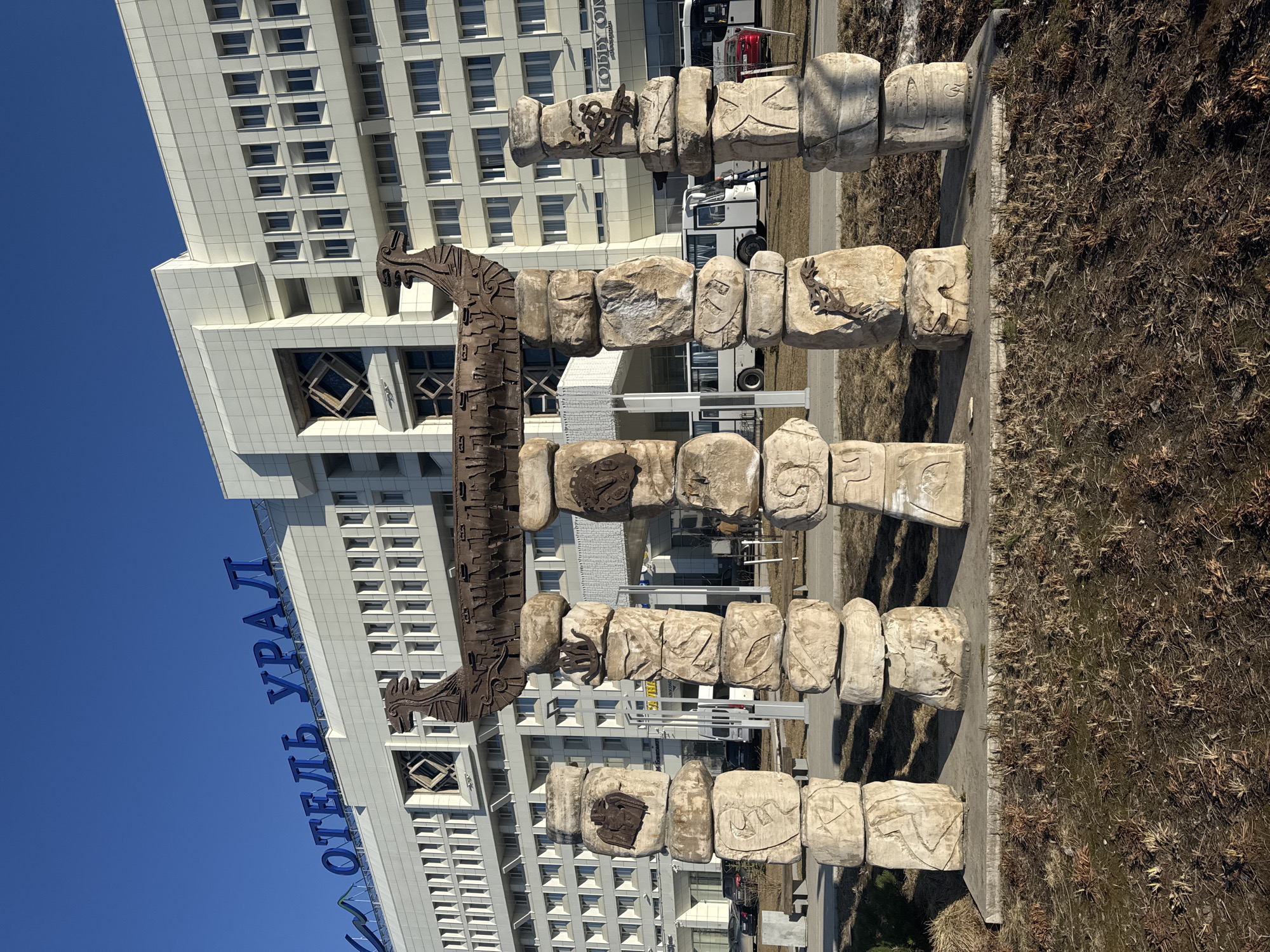 A horizontal, eye-level shot features a monument with carved stones, possibly representing ancient symbols or figures, set against the backdrop of a modern building in Perm, Russia. The monument consists of rows of roughly rectangular stone blocks, each bearing a carved image. These blocks are mounted on metal poles, creating a three-tiered structure. A decorative metal element, resembling a stylized bird or dragon, is suspended above the stone structure.
Behind the monument stands a tall, white building with multiple rows of windows. A sign in blue Cyrillic letters, presumably spelling out "Отель Урал" ("Hotel Ural"), is mounted on the building. The foreground consists of a strip of dry, brown vegetation and a concrete sidewalk. The sky is clear and blue. A white bus can be seen in the distance behind the monument. There are no people visible in the image. The overall impression is of an artistic or cultural installation located near a modern hotel in an urban environment.