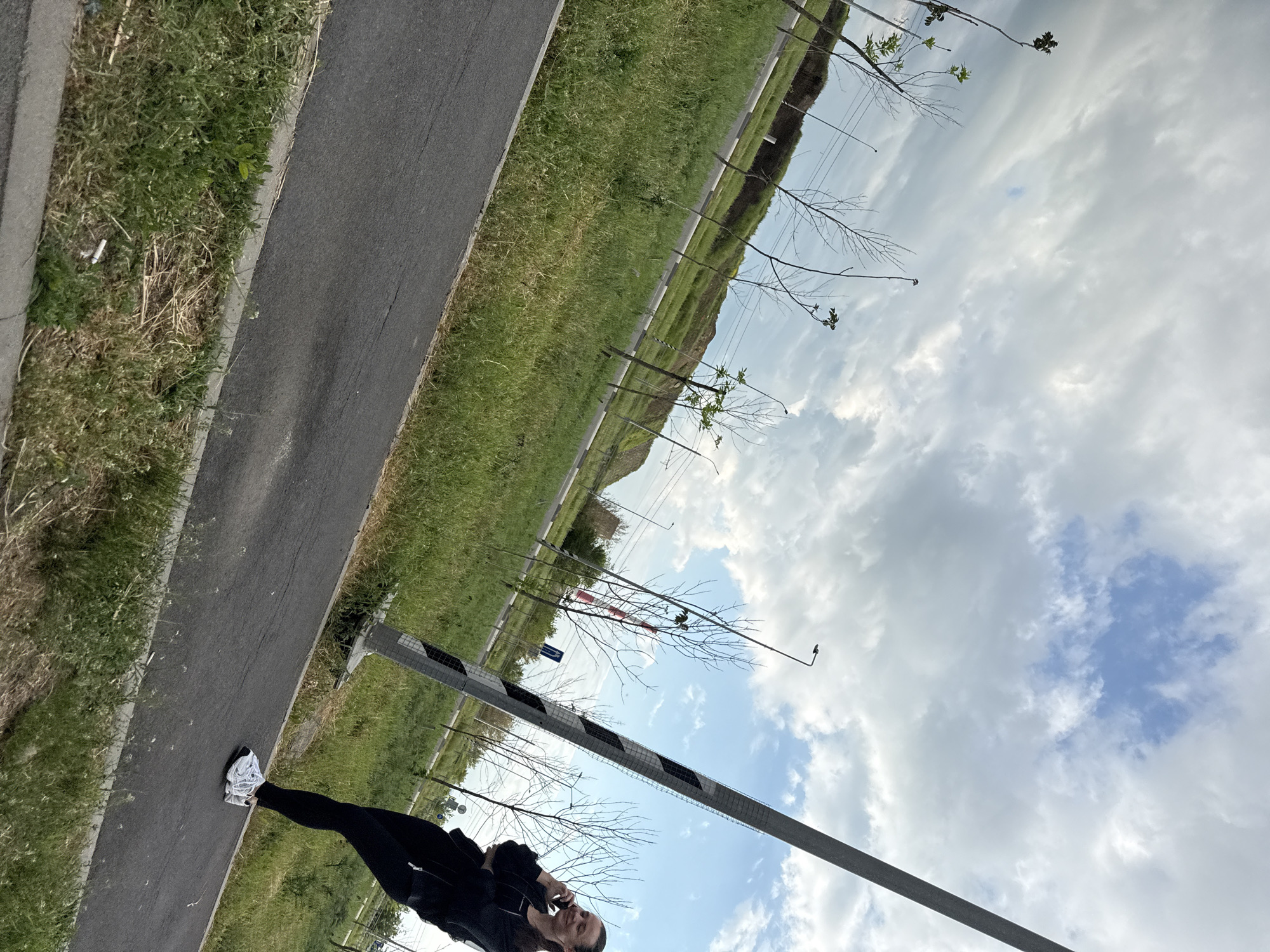 A sideways angle shot shows a woman in black clothing standing on a gray asphalt path next to green grass. The background shows sky with white clouds and some trees. A safety barrier with red and white paint stands near the woman. The location is likely near a road in Rostov-on-Don, Russia.