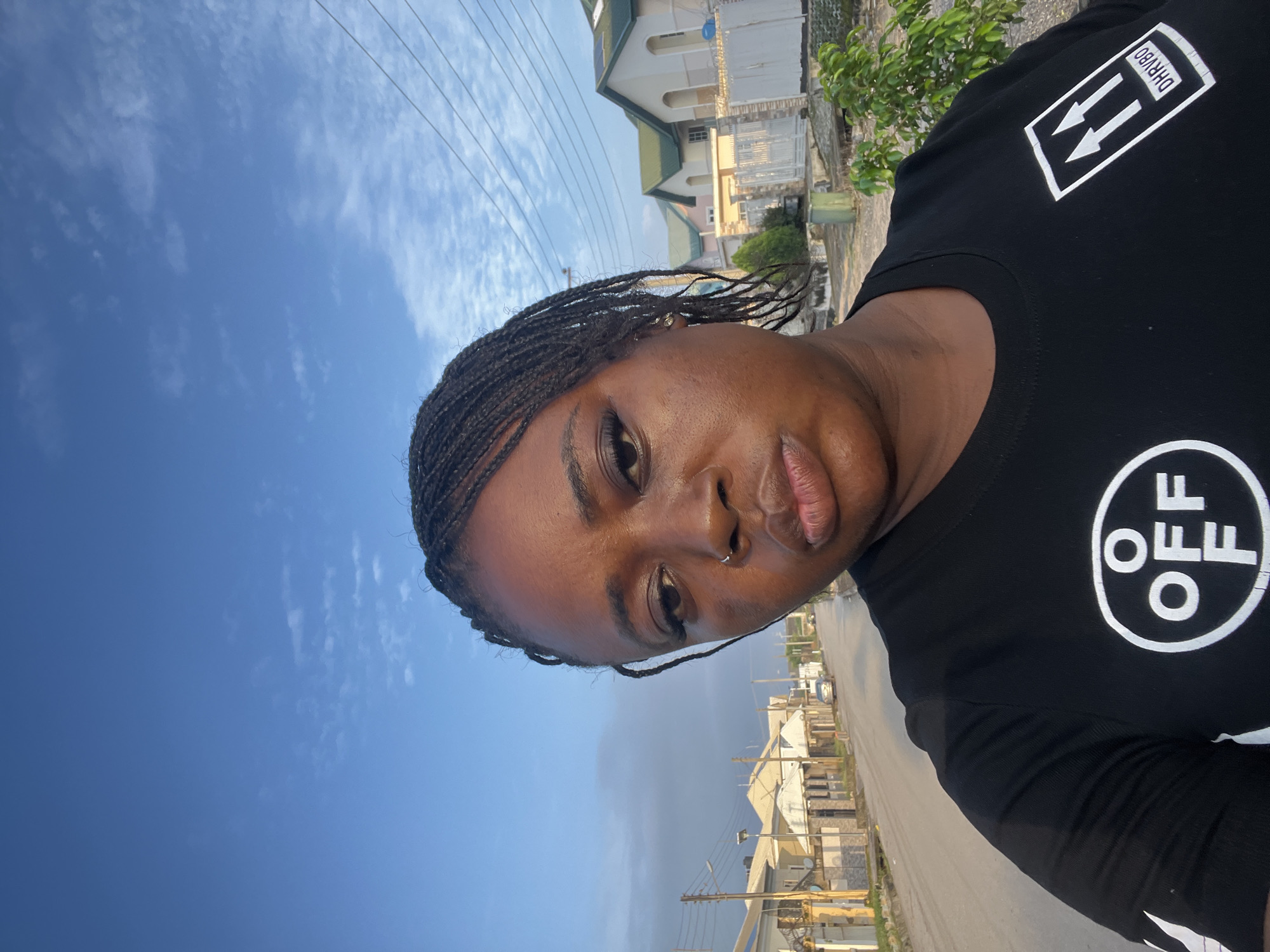 A selfie is taken outdoors in a residential area of Abuja, Nigeria. The subject, a woman with braided hair and a nose ring, is wearing a black t-shirt with white graphics. She is positioned in the foreground, with her face tilted slightly and her lips pursed. Behind her, a paved street stretches into the distance, lined with modern houses and streetlights. The sky above is a clear blue with scattered clouds. The overall impression is that of a casual snapshot taken during daylight.