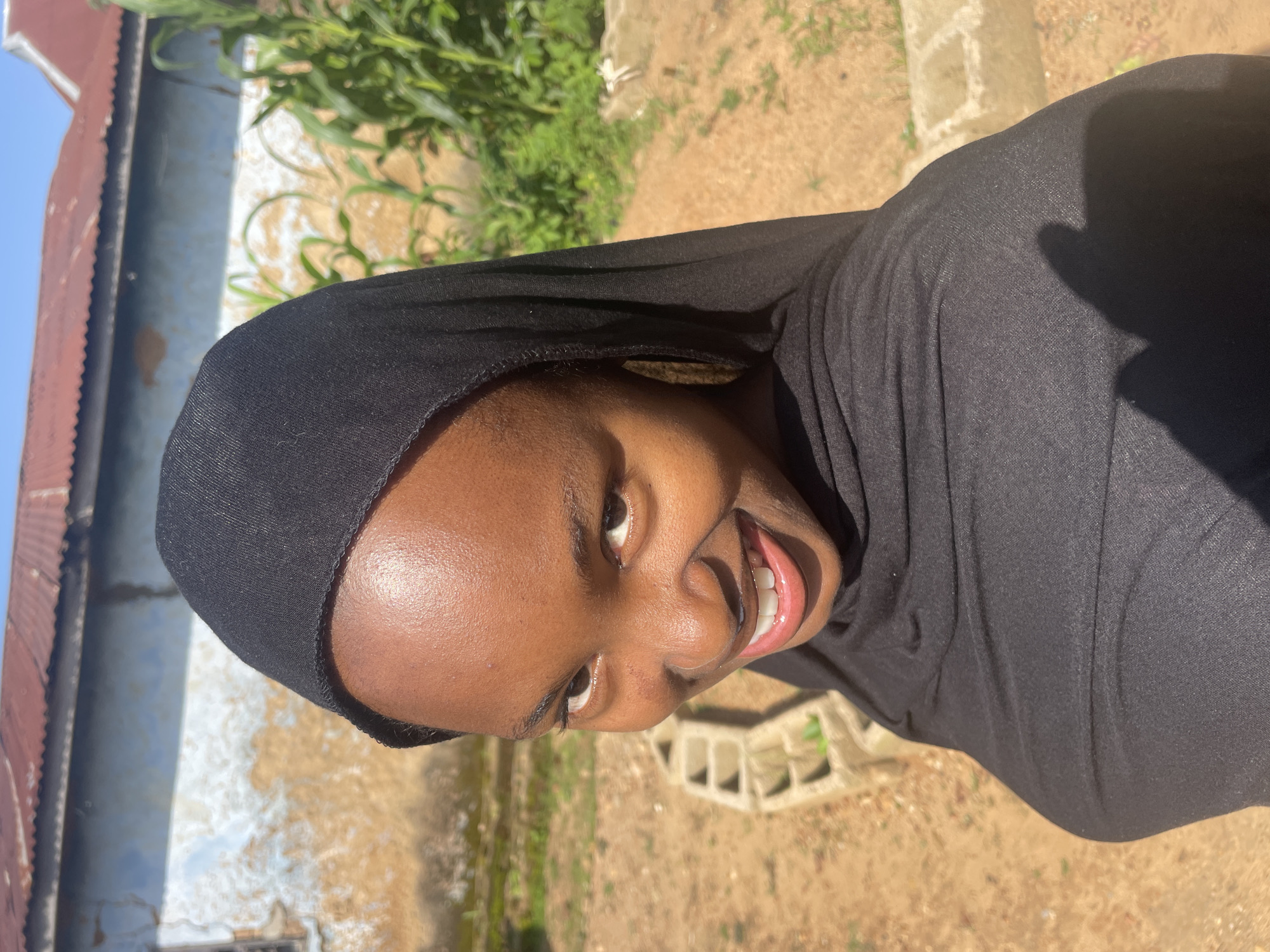 A young Black female wearing a black hijab and black garment is looking directly at the camera, smiling with her mouth slightly open, showing her teeth. She is positioned in the foreground, with her head and upper torso filling most of the frame. The background consists of a building with a corrugated metal roof and light blue and tan walls, with some vegetation to the right and what appears to be sandy ground with concrete blocks scattered around. The image appears to have been taken outdoors in daylight. The location of Jos, Nigeria, is not explicitly indicated in the image itself.