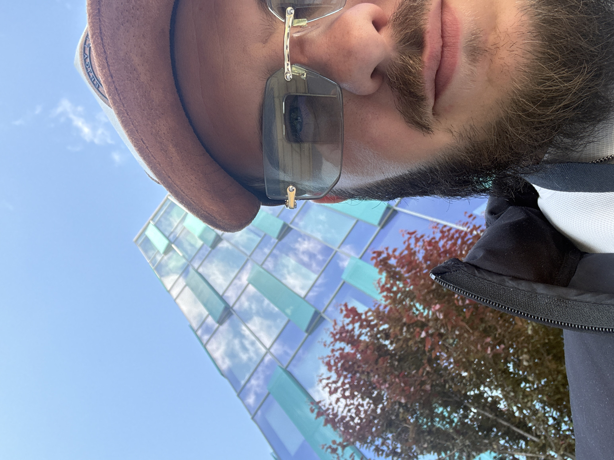 The image is a low-angle selfie of a man with brown hair, a beard, and a mustache, wearing a brown hat and rimless sunglasses with a gold-colored frame. In the background, there is a modern building with reflective windows that have blue and green panels, along with a tree with reddish-brown leaves. The sky is visible in the upper right corner and appears to be clear and blue. The man is wearing a dark-colored jacket with a zipper. The location is potentially in Sofia, Bulgaria.