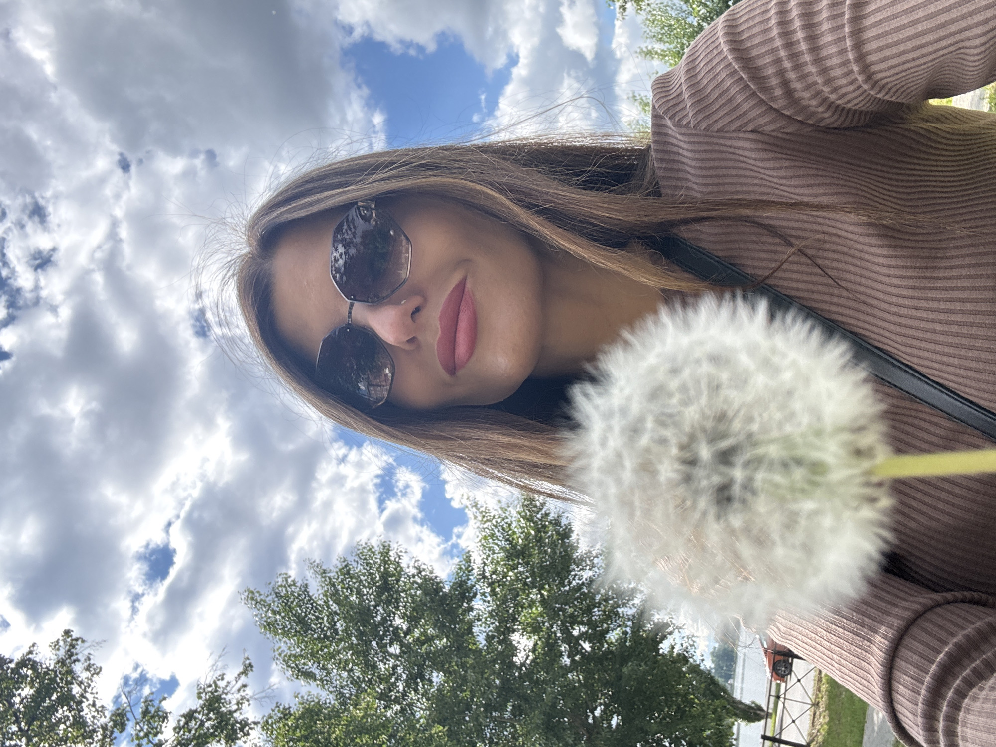 A woman is holding a dandelion in the foreground of a photograph, taken from a low-angle perspective. The woman has shoulder-length brown hair and wears sunglasses. She's wearing a light brown ribbed long-sleeved shirt. Behind her is a sky filled with white and gray clouds. In the lower left, green treetops are visible. In the distance, partially obscured by the dandelion, is what appears to be a fenced area and the edge of a body of water. Based on this description, the location is near the Votkinsk Reservoir, which could be located in the city of Votkinsk, Russia.