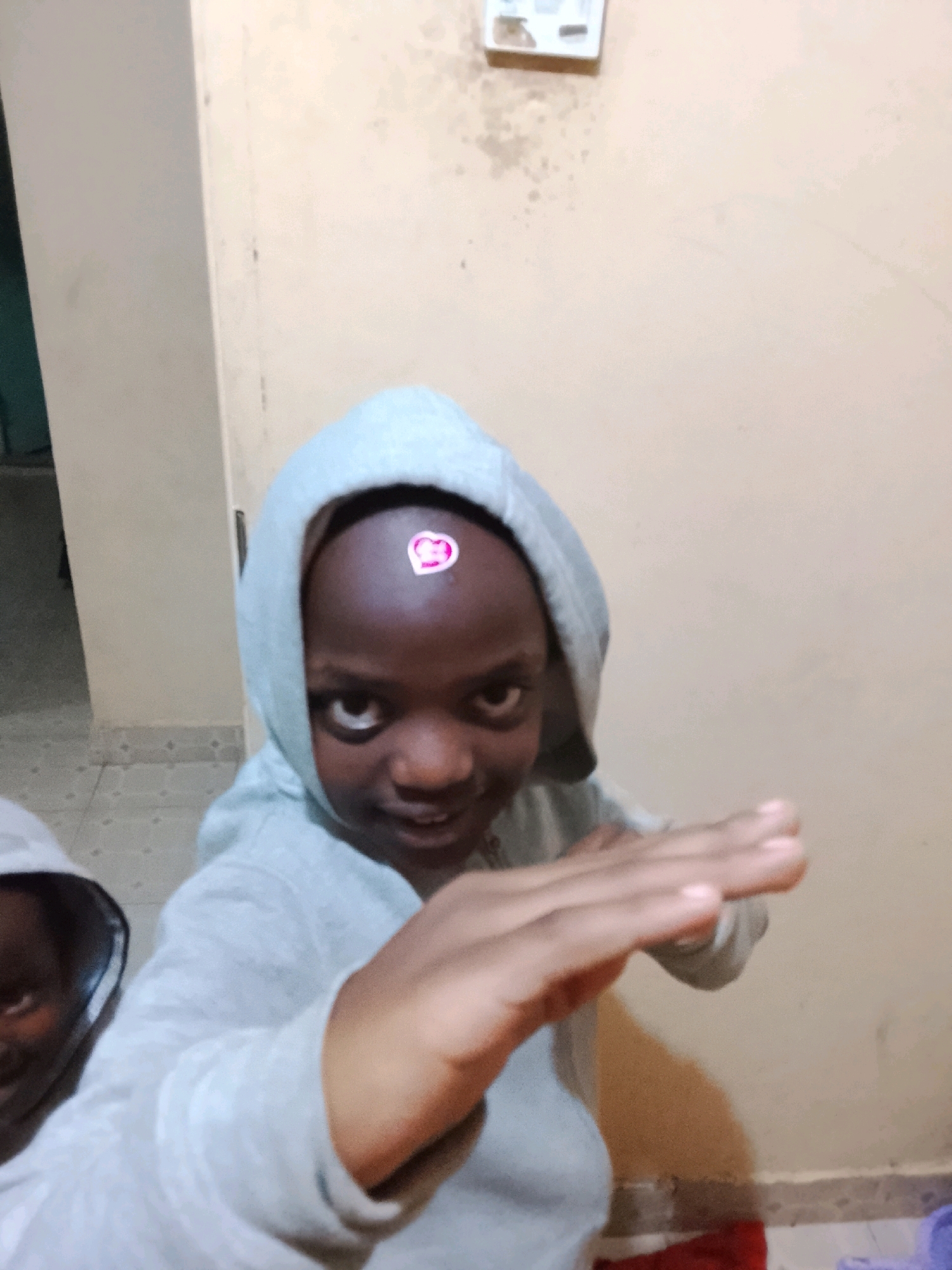 A young child, wearing a grey hooded sweatshirt, is shown from the chest up, with a sticker on their forehead. The child is looking towards the camera with a slight smile. Another child is partially visible to the left, also wearing a hooded sweatshirt. The background consists of a light-colored wall and floor, with a glimpse of a door on the left. The setting appears to be indoors, possibly a home or a communal space. The image is taken in the city of Unknown, Unknown.