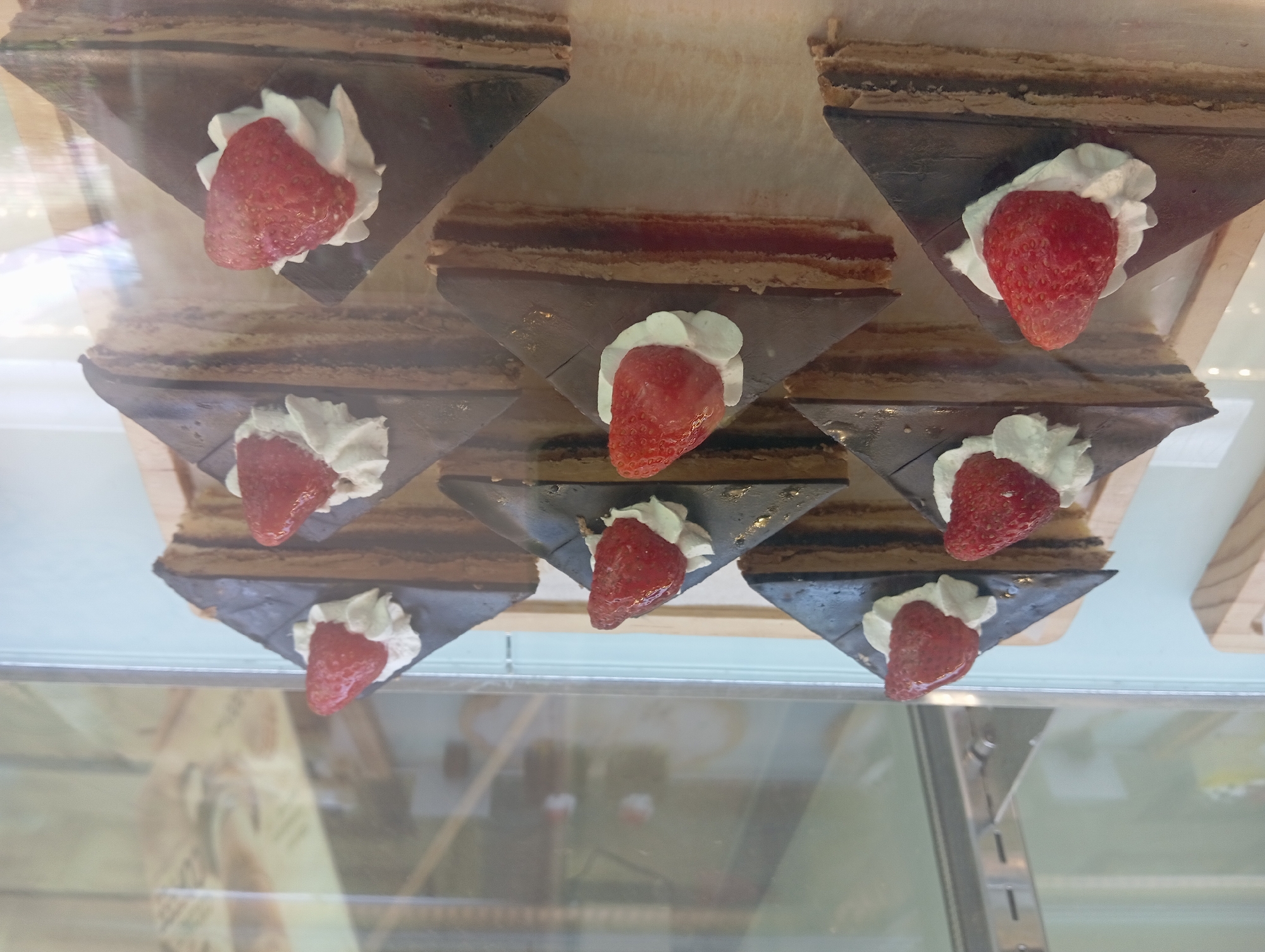 A display case in a Nha Trang, Vietnam bakery showcases eight triangular chocolate cakes, each topped with whipped cream and a fresh strawberry. The cakes are arranged on a light-colored wooden tray. The display case has a glass front, reflecting the interior of the bakery, including shelves and other baked goods. The lighting in the scene is bright and artificial.