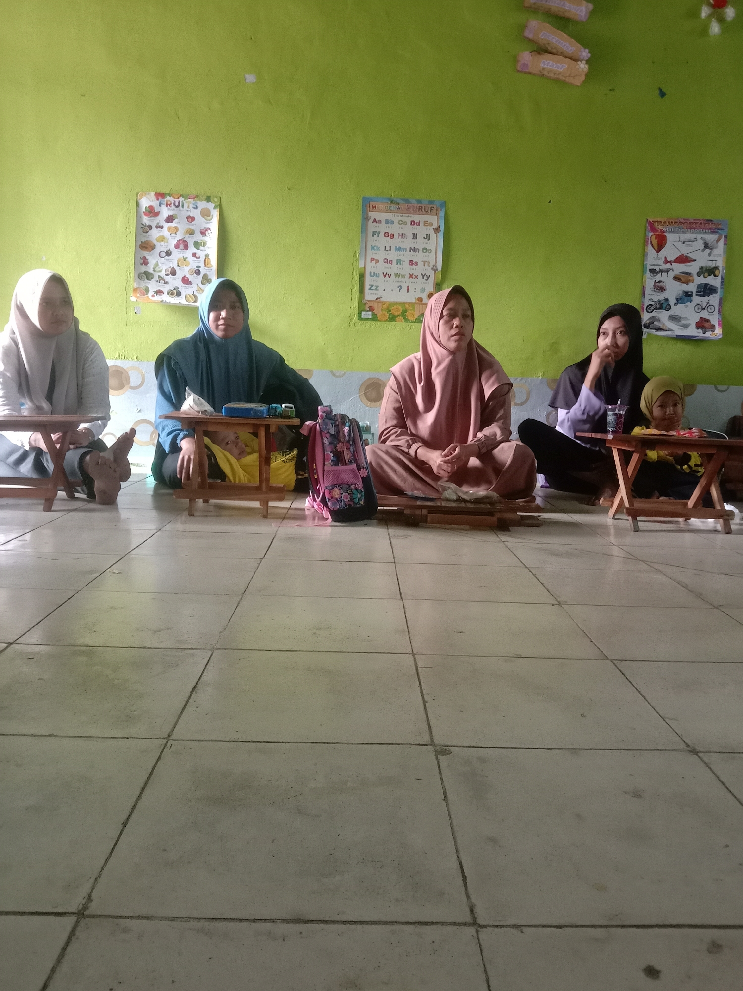 In Sambongrejo, Indonesia, a group of five individuals are seated on a tiled floor in front of a pale green wall adorned with educational posters. On the left, two people wearing hijabs are seated behind small wooden tables. The person on the far left wears a light grey hijab and a grey garment. The person next to them wears a dark blue hijab and a matching outfit. Between them and the next seated group, a child in a yellow hijab is partially visible, peeking out from under a wooden table with a pink and black backpack beside it.
To the right, two individuals are seated. The first, wearing a light brown hijab and a matching outfit, is looking towards their right. The second, wearing a black hijab and a purple top, is looking towards the center of the scene. Next to the person in the black hijab is a young child wearing a yellow hijab and a black and yellow shirt, seated behind a low wooden table. They are holding a glass with a straw. Educational posters depicting fruits, the alphabet, and vehicles are visible on the green wall behind the seated individuals. The floor is covered with light-colored square tiles.