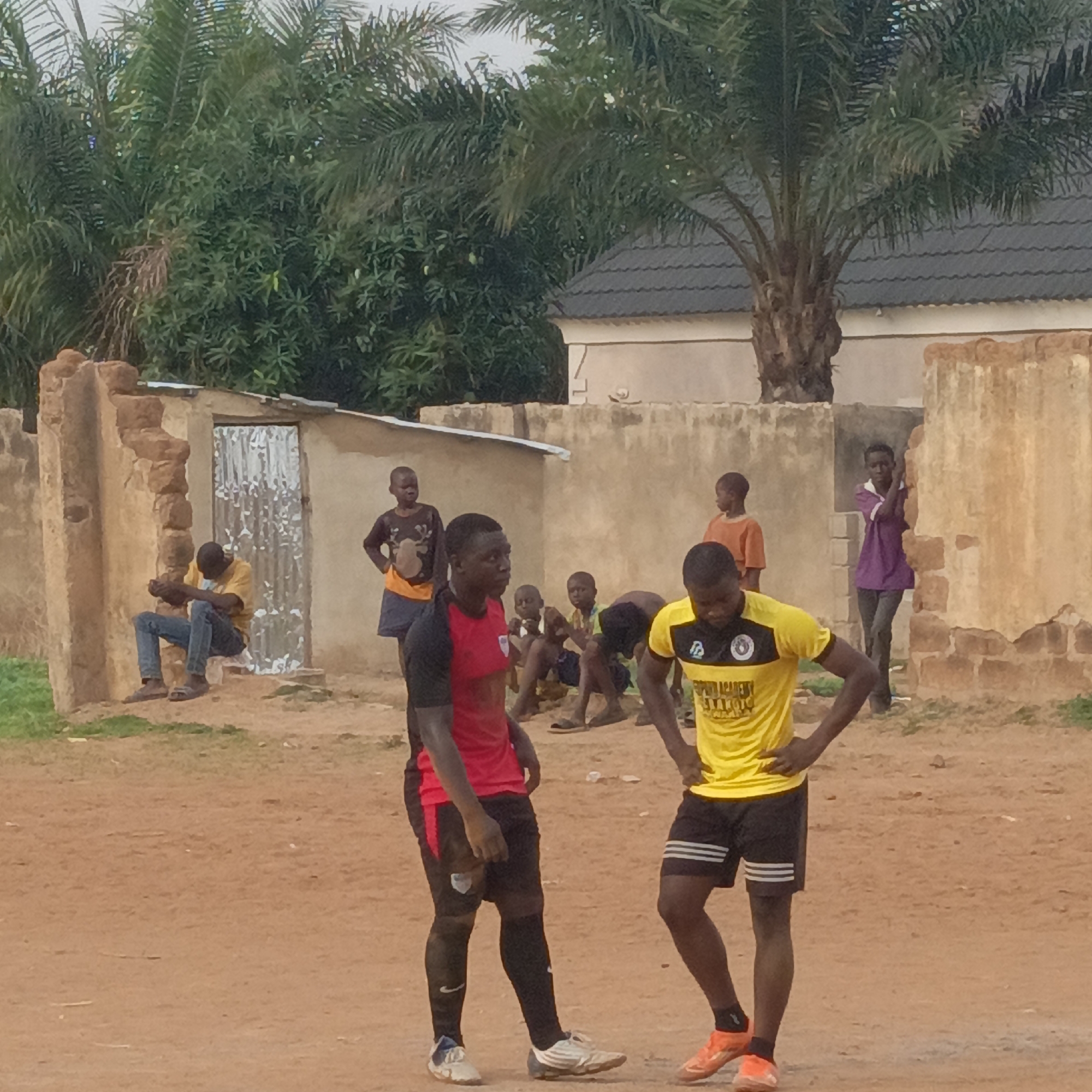 This image captures an outdoor scene in Akwanga, Nigeria, depicting a group of people, primarily young males, in what appears to be a casual or informal gathering.
In the foreground, two young men stand on reddish-brown dirt ground, both dressed in soccer attire. The man on the left wears a red and black short-sleeved jersey, black shorts, and black long socks with a white Nike swoosh, complemented by white and blue athletic shoes. The man on the right is in a yellow and black short-sleeved jersey that displays "SPORTS ACADEMY" and partially visible text "WARRIORS," along with a crest on the chest. He wears black shorts with white stripes and orange athletic shoes. Both men have their hands on their hips, suggesting they are resting or observing.
In the mid-ground, several other individuals are scattered. To the left, a man in a yellow shirt and blue jeans sits on a low, weathered wall next to a structure with a corrugated metal door. Further to the right, a small group of children and young adults are seen standing or sitting near additional low, earthen-colored walls, which appear aged or unfinished. One child wears an orange shirt, and another in the background wears a purple shirt, leaning against a wall.
The setting features simple, rustic structures made of what appears to be mud bricks or similar materials, some showing signs of disrepair. Lush green vegetation, including prominent palm trees, forms a dense background, with a building featuring a dark tiled roof partially visible amidst the foliage on the right side. The ground is unpaved and dusty. The sky is overcast, providing soft, diffused lighting, indicating either early morning, late afternoon, or a heavily clouded day.