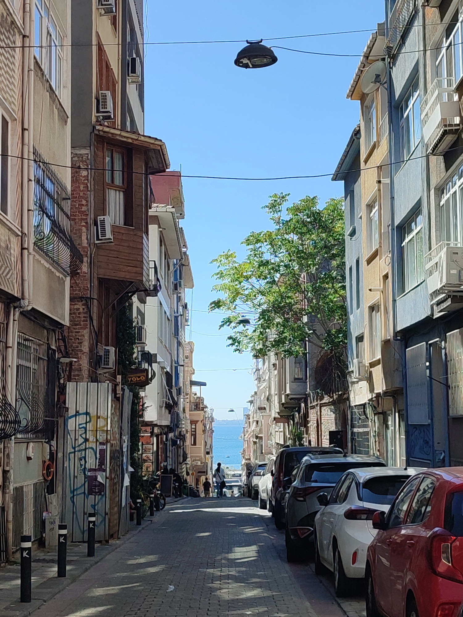The image depicts a narrow street in the Kadıköy district of Istanbul, Turkey, leading towards the sea. The street is paved with cobblestones and lined with multi-story buildings on both sides. The architecture is varied, with older brick buildings interspersed with more modern structures, many featuring balconies, air conditioning units, and satellite dishes. On the left side, a corrugated metal fence with graffiti is visible. On the right, several cars are parked along the street, including a red car in the foreground. Two figures, a man and a child, are walking away from the camera further up the street, heading towards the water. A large green tree grows between buildings on the left side, its branches reaching towards the sky. Overhead, electrical wires and a street lamp are strung across the street. The sky is clear and blue, and the sea is visible in the distance. The scene suggests a sunny day in a residential or mixed-use urban area.