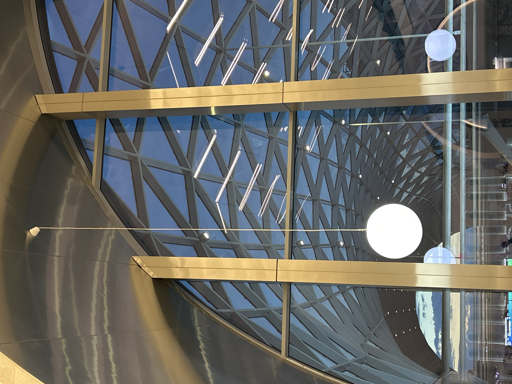 The image displays the interior of a large, modern building, characterized by its expansive glass walls and metallic structural elements. The perspective is tilted, showcasing a tall, curved glass section framed by a complex, geometric grid of light-colored metal beams. Through the glass, a clear blue sky with soft cloud reflections is visible, indicating it is daytime.
Two prominent, thick horizontal beams, appearing to be made of gold-toned or brushed metallic material, cut across the middle ground, serving as a significant architectural feature. On the left side, a curved, reflective wall, possibly made of brushed aluminum or steel, runs vertically, reflecting light in shimmering streaks.
The scene is filled with reflections of various light sources. Numerous linear, bright white lights, resembling LED strips or fluorescent tubes, are clearly reflected across the glass panels, creating a sense of depth and artificial illumination. A large, spherical or disc-shaped white light fixture hangs by a thin wire prominently in the center-right of the frame, with a smaller, similar fixture visible further in the background. Another small, dome-shaped light is partially reflected near the main circular fixture.
The overall impression is one of vastness and contemporary design, typical of a modern airport, convention center, or a large public atrium. The complex interplay of natural light, artificial illumination, and reflections creates a dynamic visual. No people or specific activities are visible, emphasizing the architectural grandeur of the space. There is no visible text.