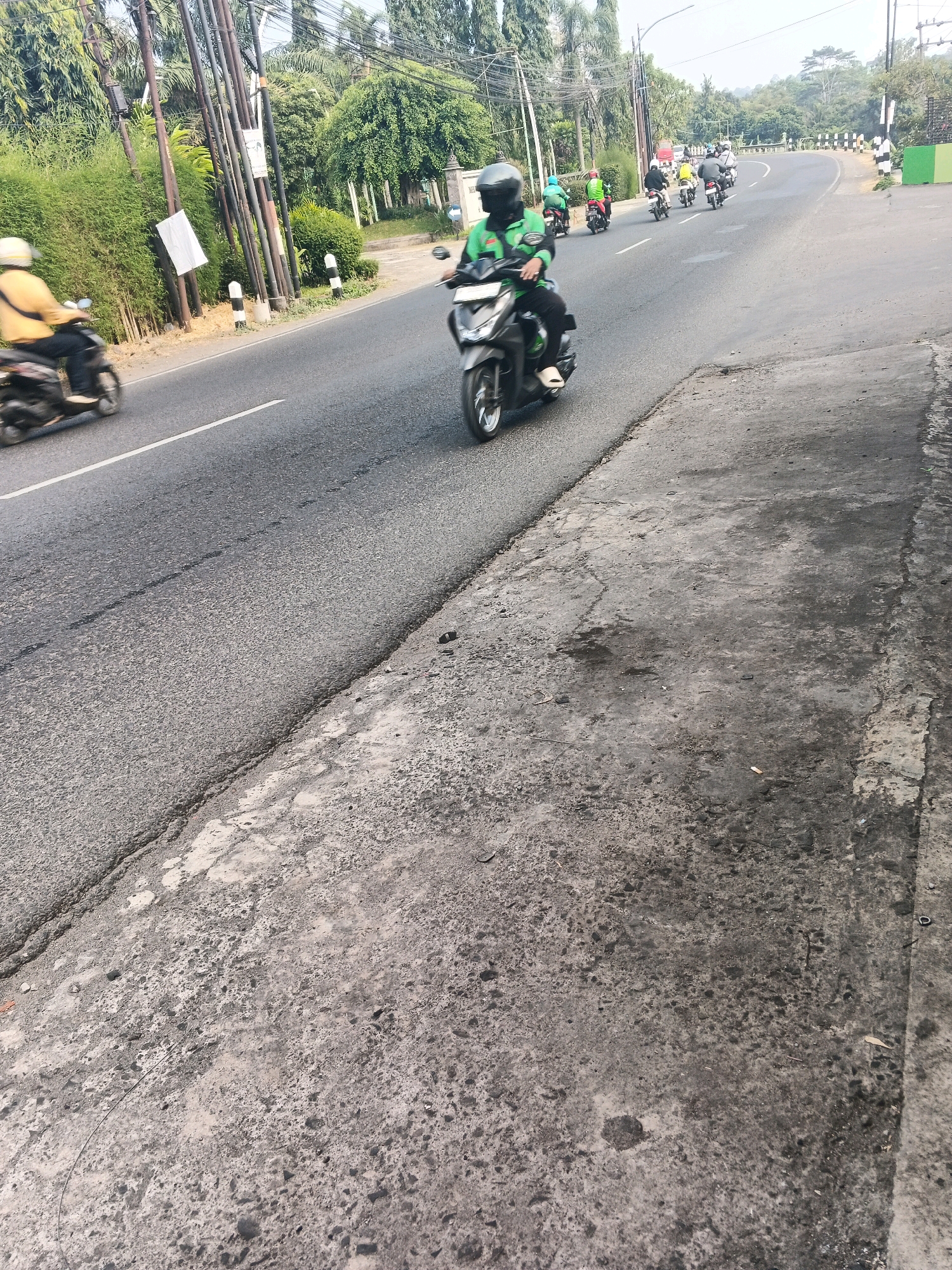 This is an eye-level, medium shot taken from the side of a two-lane road in Ungaran, Indonesia. The road appears to be paved and has a white dividing line. The scene is populated by multiple people riding motorcycles, predominantly in the middle and right lanes, moving away from the camera. A person wearing a green jacket and helmet is the most prominent subject, riding a dark-colored motorcycle in the foreground, left lane. To the left of the road, there is a paved shoulder, then a dirt and gravel area, followed by a dense green bush and bamboo hedge, and further left, utility poles and cables. In the background, the road curves to the right, with more motorcycles in the distance. The environment is outdoors with a slightly overcast sky, and lush green vegetation and trees line the road, suggesting a semi-urban or rural setting on the outskirts of Ungaran.