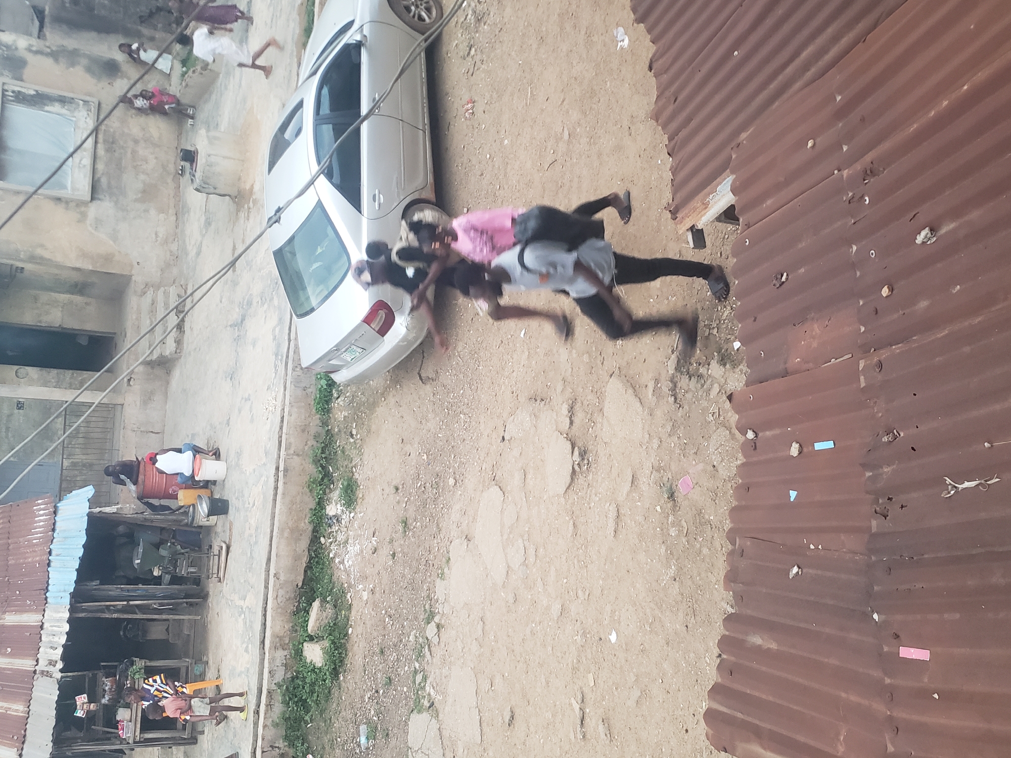 The image shows an outdoor scene in Alalubosa, Nigeria. A silver sedan is parked on a dirt surface. Several people are present, including a group of children on the left and two men engaged in what appears to be a physical activity, with one man carrying the other on his back. Buildings with corrugated metal roofing are visible on either side of the dirt area. There are additional people and objects in the background, suggesting a busy, densely populated environment.