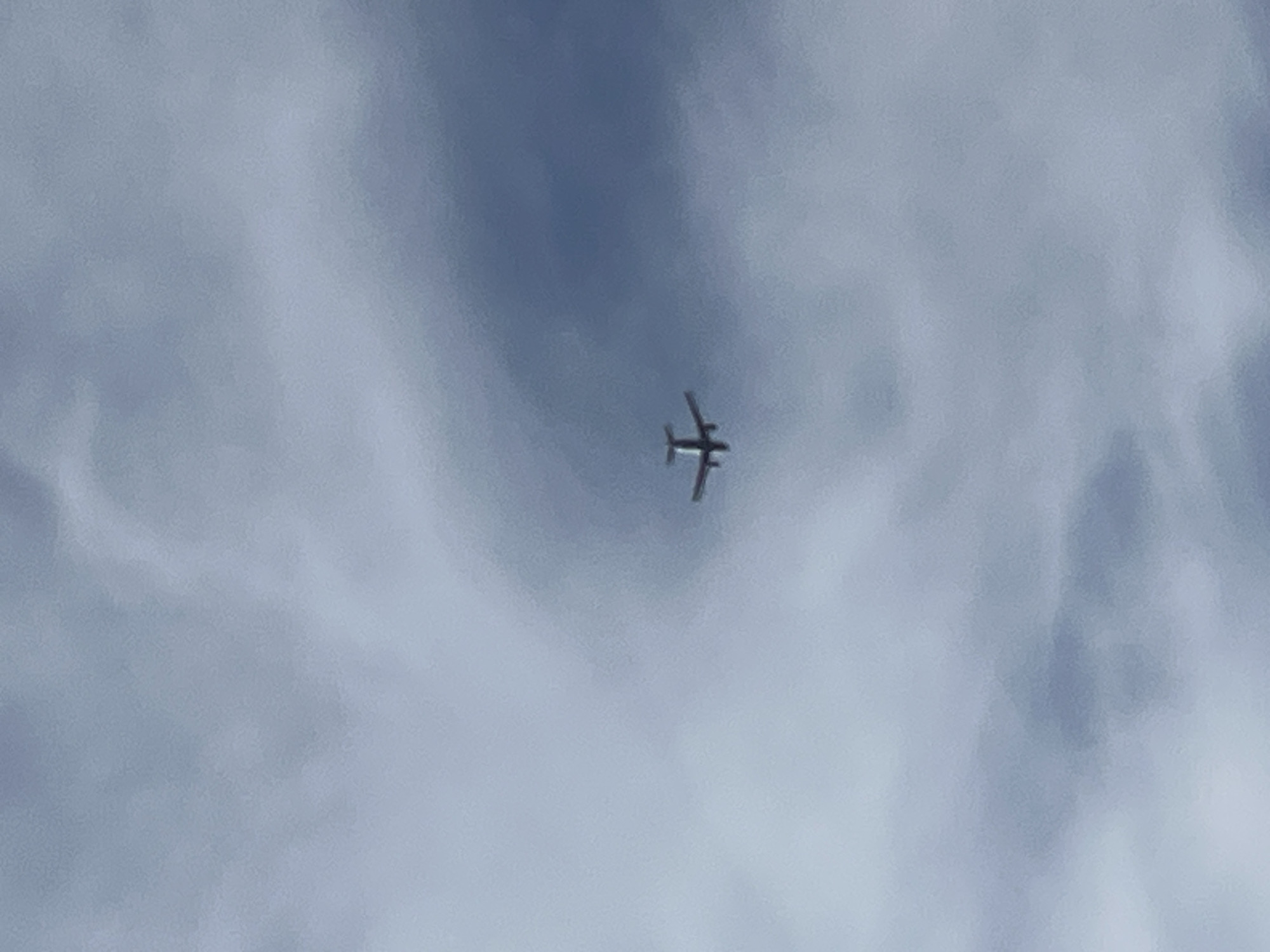 A small, twin-engine airplane is visible in the sky, flying from left to right. The plane appears to be at a moderate altitude against a cloudy, overcast sky. The clouds are a mix of white and grey, obscuring the sun and creating a diffused light. The overall scene is muted in color, dominated by shades of blue and grey. The location is Prijevor, Montenegro.