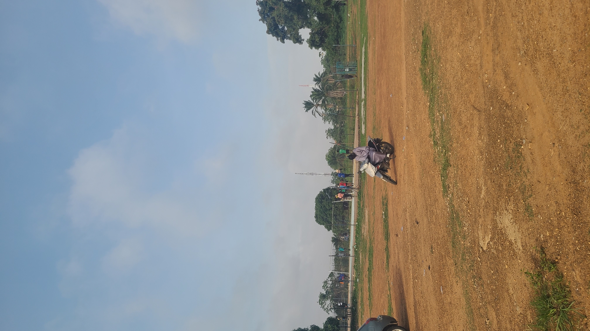 The image depicts an outdoor scene in Ogbomosho, Nigeria, featuring a dirt field bordered by trees and a fence. A motorcycle is parked on the field, with a person seated on it. Several other people are standing nearby. A communication tower is visible in the background. A basketball hoop stands to the left of the field. The sky is light blue with scattered clouds. Part of a car is seen at the bottom of the frame.