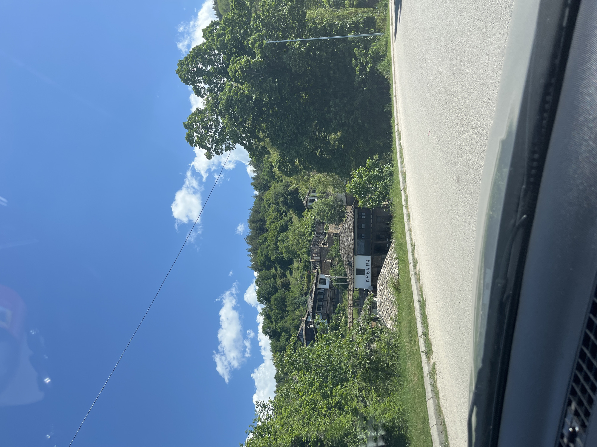 The image captures a view from inside a vehicle traveling along a road in Leshten, Bulgaria. On the roadside, a traditional Bulgarian building, possibly a guesthouse, is nestled on a hillside amidst dense greenery. A sign displaying the Cyrillic inscription "КРЪЧМА" is visible next to the building. The sky above is blue with scattered cumulus clouds. An overhead electrical line stretches diagonally across the sky. The road appears to be paved and well-maintained, running parallel to the vegetated hillside.