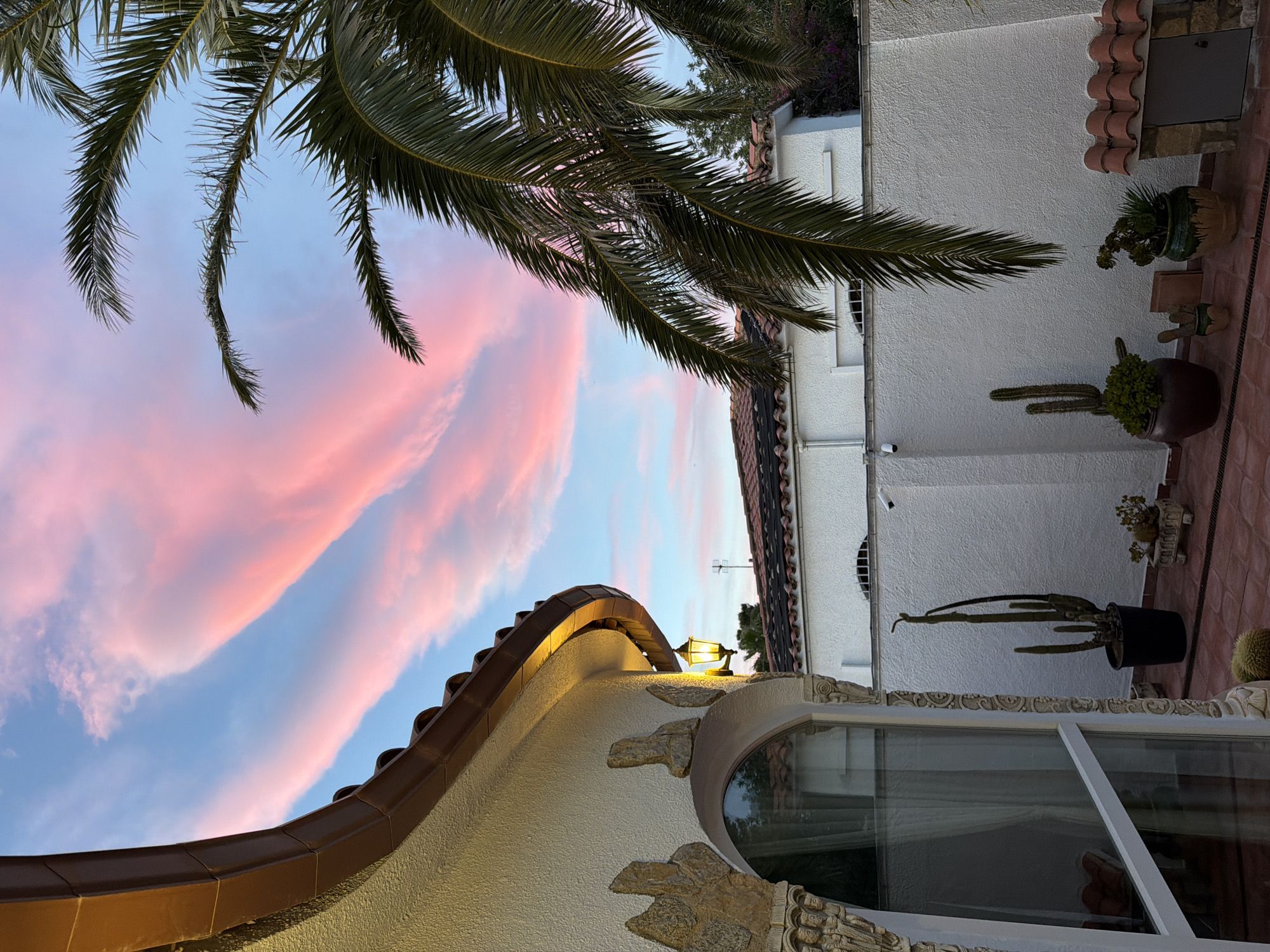 The image depicts an exterior scene in Mont-roig del Camp, Spain, at dusk or dawn, with a focus on architectural elements and a colorful sky. A palm tree frond dominates the upper left portion of the frame. A building with a curved roofline and textured exterior wall is visible in the foreground, illuminated by an exterior light fixture. A window with decorative stonework surrounds it. In the background, a white building with a tiled roof and various potted plants and cacti are arranged on a tiled patio. The sky is a gradient of blue and pink, suggesting either sunrise or sunset. The composition is shot from a low angle, looking upwards.