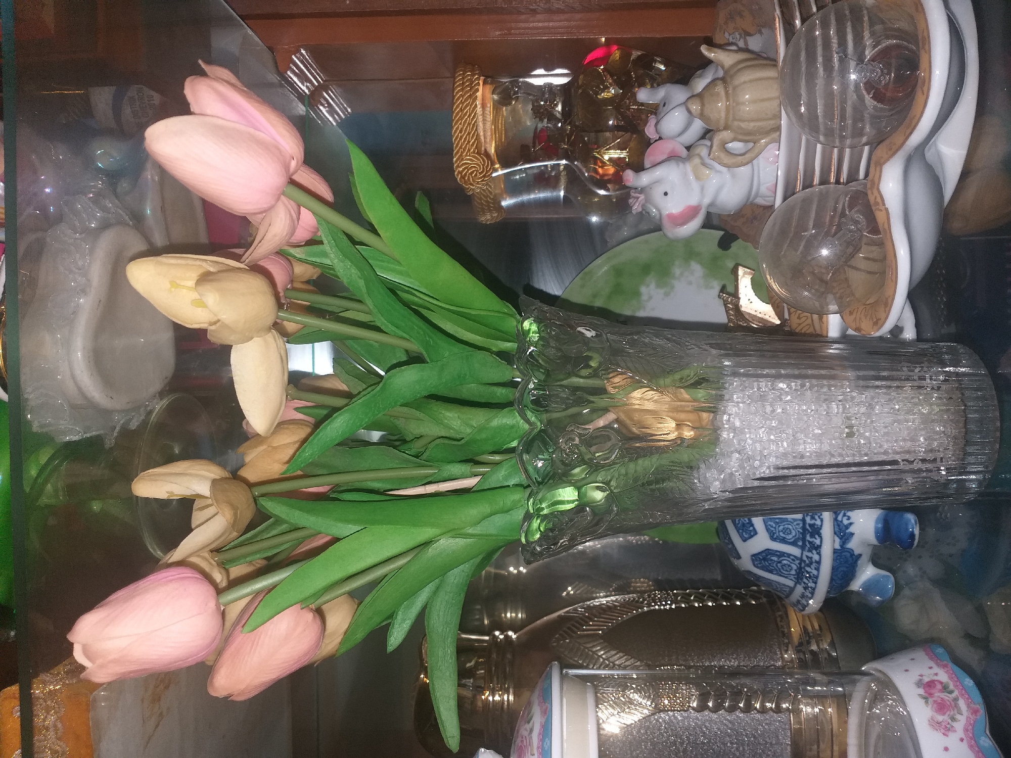 The image shows a collection of decorative items on a glass shelf. In the foreground, a clear glass vase is filled with clear crystals and artificial tulips with pink and light yellow petals. To the left of the vase, a white and blue ceramic turtle figurine is visible. Further to the left, a portion of a decorative container with floral patterns is present. To the right of the tulip vase, there are two decorative glass globes on a white and gold decorative dish, each containing a light bulb. Behind these globes are two white ceramic elephant figurines, one with pink ears and a yellow bow, and a small ceramic teapot. Another decorative glass bottle with a gold rope detail is positioned behind the elephant figurines. The background shows more decorative items and a wooden cabinet. The scene does not provide any information about the city of Dumai, Indonesia.