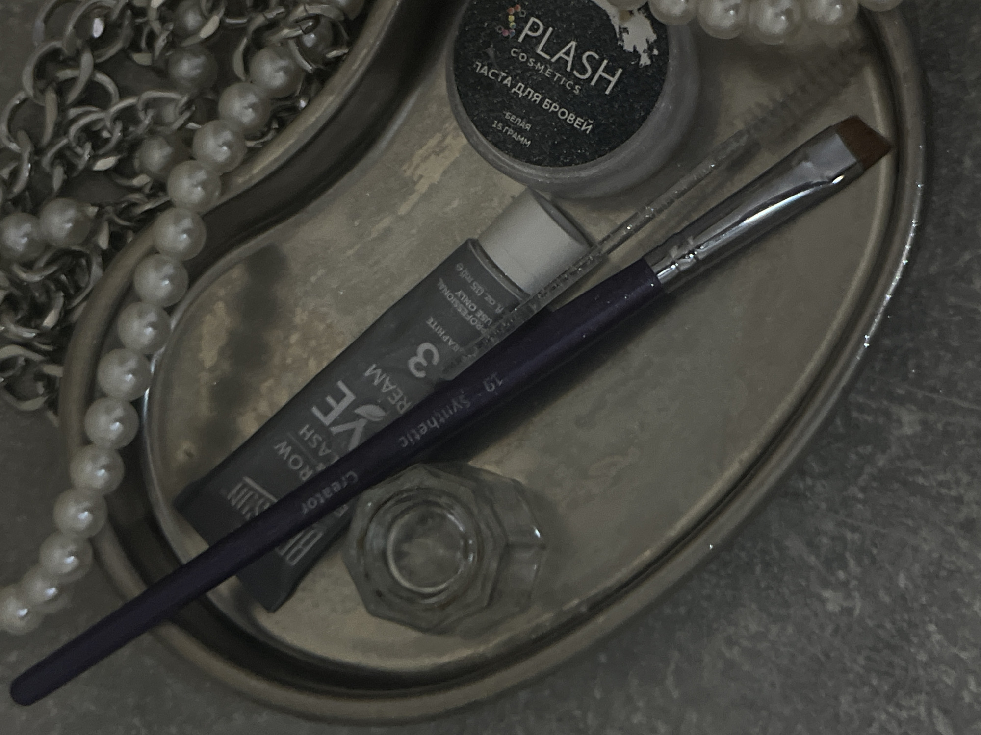 This close-up, overhead shot shows a collection of cosmetic items arranged on a metallic tray. The tray itself appears to be kidney-shaped, with a smooth, reflective surface. To the left, a strand of faux pearls is draped, interspersed with a chunky silver chain.
The focal point of the image is a set of eyebrow grooming tools. A small, dark jar with a label that reads "PLASH COSMETICS ПАСТА ДЛЯ БРОВЕЙ БЕЛАЯ 15 ΓΡΑΜΜ" is visible at the top center. Below this jar is a tube of what appears to be brow cream or gel, with text indicating "PROFESSIONAL USE ONLY fl oz. (15 ml e)" and "GRAPHITE". A cosmetic brush with a purple handle and a flat, angled synthetic head is placed diagonally across the tray, positioned over the brow cream tube. Next to the tube is a small, hexagonal glass cap or container.
The lighting is dim, creating a somewhat moody and luxurious atmosphere. The metallic sheen of the tray and jewelry contrasts with the matte textures of the cosmetic products. The overall scene suggests a personal grooming or makeup application setup, possibly in a bathroom or dressing area. The Russian text on the jar indicates a potential location in Russia. No people are visible in the image.