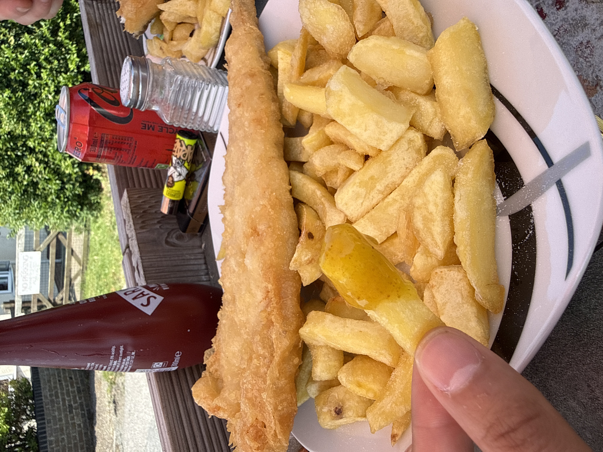 The image shows a plate of fish and chips, a bottle of tomato sauce, and a can of Coca-Cola on a wooden table. A salt shaker and a lighter are also visible on the table. The background shows a brick wall, a wooden gate, and greenery. The scene appears to be outdoors, possibly at a restaurant or cafe in Great Yarmouth, United Kingdom, given the context provided. The lighting suggests it is daytime. A person's hand is holding one of the chips. The food looks freshly prepared and the overall scene depicts a casual meal.