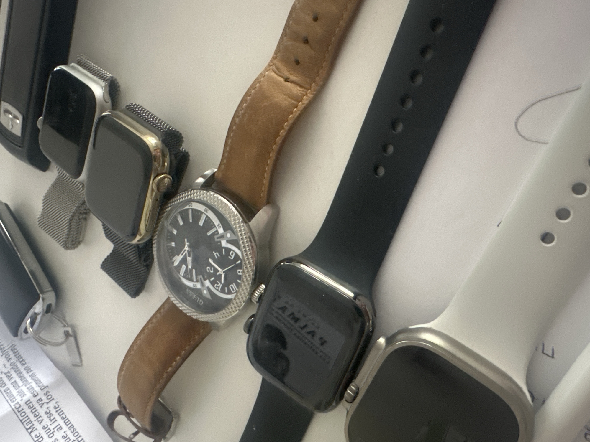 The image shows a collection of wristwatches on a light surface, likely a table. There are several smartwatches with different band colors (black, white, silver mesh), and a traditional analog watch with a brown leather strap. The analog watch has a silver casing and a black face with numerals. Text in a document is partially visible in the bottom left corner, and includes the phrase "de Mallorca existen". The text "GUESS" is visible on the analog watch. The text AMJAY is visible on the smartwatch with the black strap. There is nothing in the image indicating this location is in Sofia, Bulgaria.
