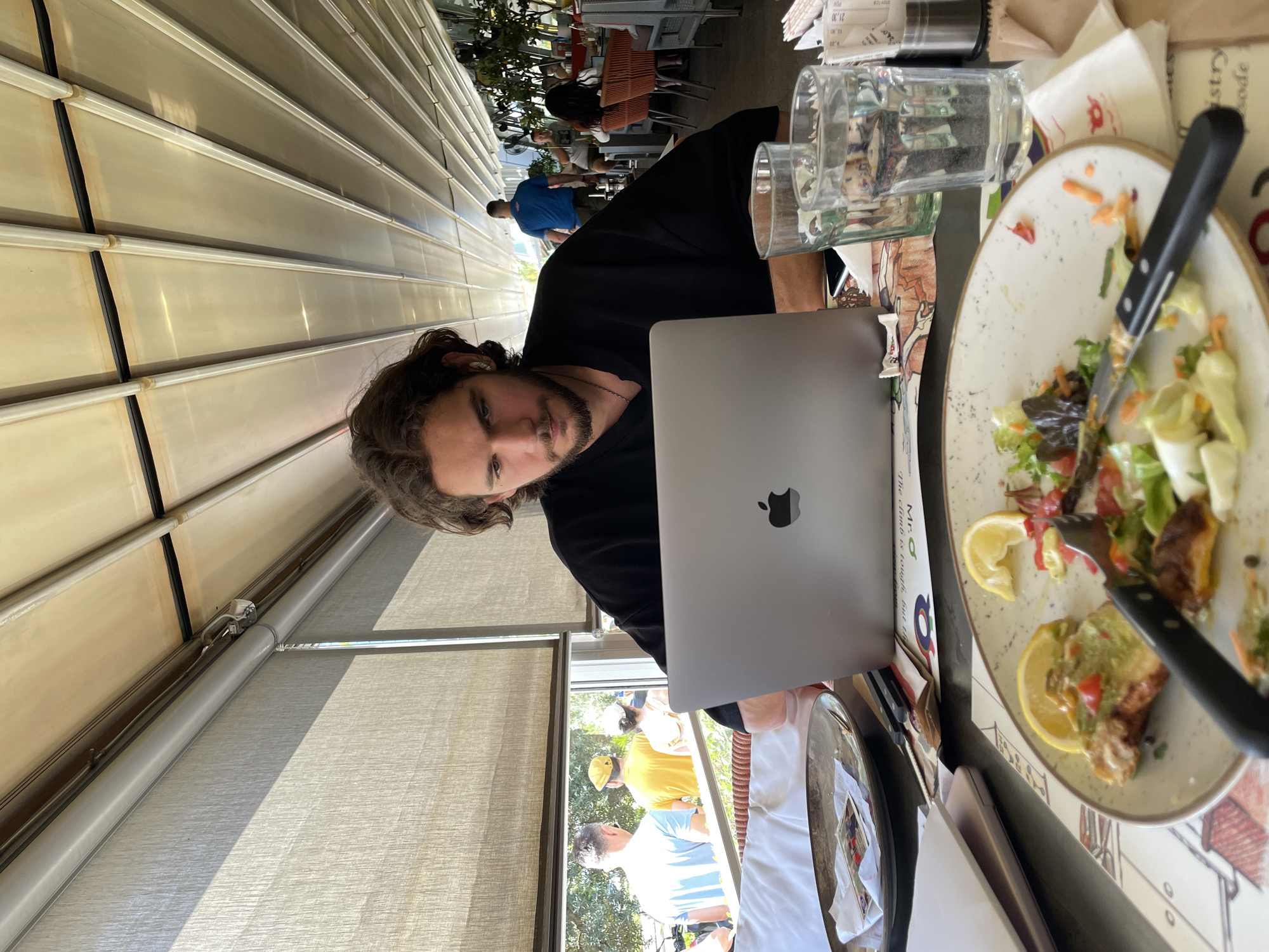 A man with dark, curly hair is sitting at a table outdoors, looking down at a silver laptop. He is wearing a black, loose-fitting shirt. The table in front of him has a partially eaten meal on a plate, cutlery, two glasses of water, and a stack of papers. In the background, through a window, other people are visible walking and sitting at tables in what appears to be an outdoor restaurant. The setting is an open-air restaurant, likely with a retractable roof, as indicated by the ribbed ceiling structure. The restaurant is situated in Budva, Montenegro, given the context.