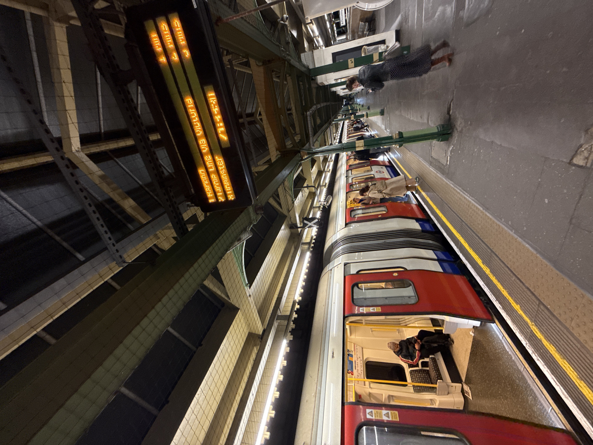 The image shows a London Underground station platform, likely in central London, UK. A train is at the platform with passengers both inside the train and waiting on the platform. A digital display shows train information. The station structure has visible beams and support structures. A few individuals are visible on the platform, some waiting and one walking towards the camera. Inside the train, passengers are seated.