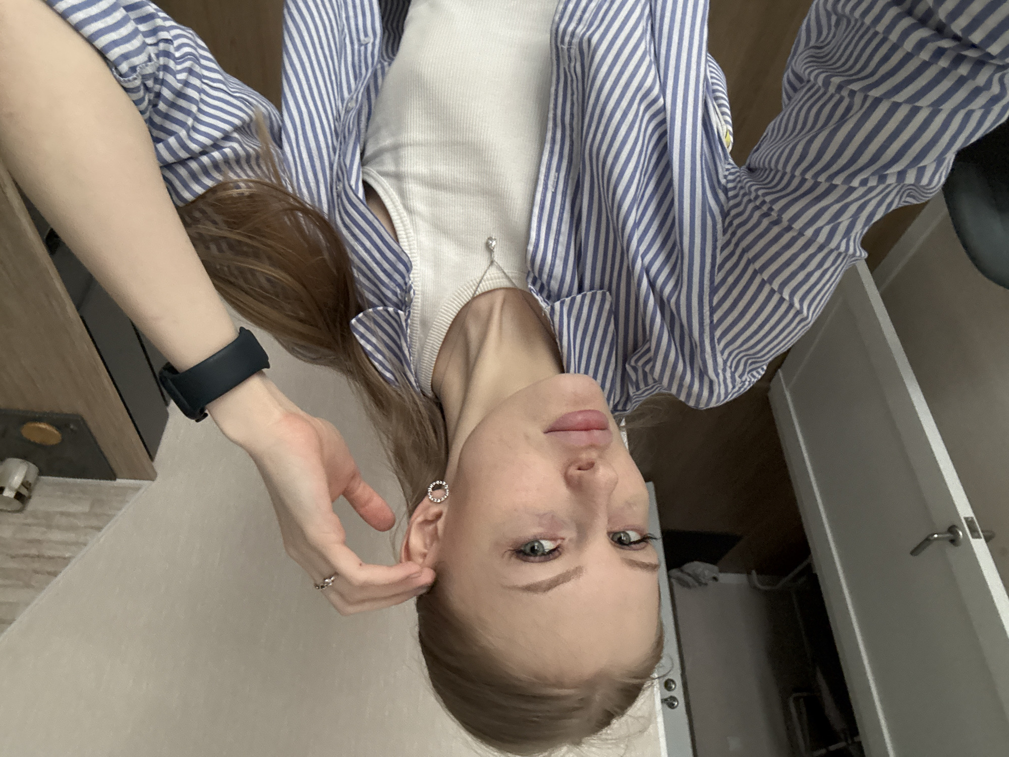 The image captures a selfie of a young individual, likely female, within an indoor setting. The photo is taken from an upside-down perspective, possibly indicating how the phone was held.
**Subjects:**
The primary subject is a person with fair skin, light blue-green eyes, and long, light brown hair styled down. They are wearing a casual outfit consisting of a white ribbed tank top layered under a blue and white striped long-sleeved button-down shirt, left unbuttoned. Accessories include a delicate silver necklace with a small pendant, a silver ring on the left ring finger, and circular silver stud earrings embellished with small stones. A black fitness tracker or smartwatch is visible on their left wrist.
**Activity or event taking place:**
The person is taking a selfie, with one hand gently touching the area near their left ear, possibly adjusting hair or an earring, while maintaining a direct gaze towards the camera.
**Notable details:**
The setting appears to be a modern room, possibly an apartment in Perm, Russia, characterized by light-colored walls, a light-toned wooden or laminate floor, and white interior doors with silver handles. A dark piece of furniture is partially visible in the background. The lighting is soft and even, suggesting daytime indoors. The person's expression is neutral and calm. No visible text is present in the image.