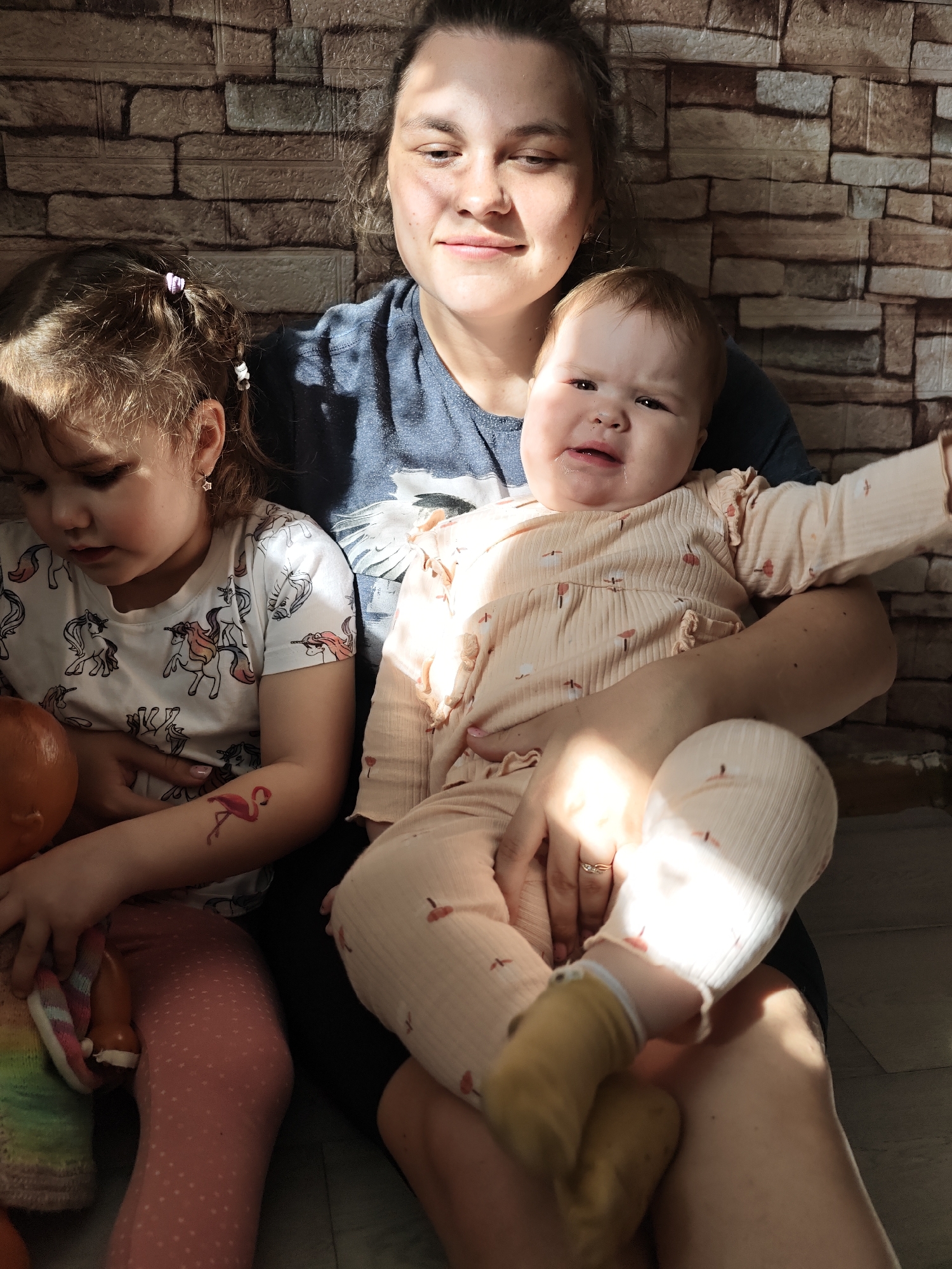The image shows a woman sitting with two young children. The woman is fair-skinned with dark hair pulled back and wears a blue t-shirt. She is holding a baby who is fair-skinned with reddish hair and wearing a light pink outfit with small bird patterns. The baby appears to be crying. To the left of the woman is a slightly older child who is also fair-skinned with light brown hair pulled back with white clips. The child wears a white t-shirt with unicorn designs and pink leggings with polka dots. She is holding a doll and appears to be looking down at it. A red flamingo tattoo is visible on the child's arm. The background is a textured brick wall. The light is bright and appears to be coming from the side, casting shadows across the scene. Based on the geo-location tag, the location is Уральское, Russia.