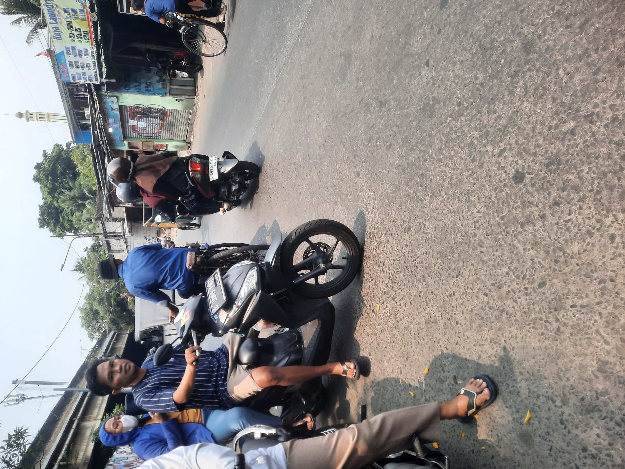 The image depicts a street scene in North Jakarta, Indonesia, likely in the Tanjung Priok area due to the presence of a mosque minaret visible in the background and the general urban, somewhat industrial aesthetic. The foreground shows several people on motorcycles. A man in a striped blue shirt and grey shorts is riding a black scooter, with a woman in a blue jacket and white mask as his passenger. Further back, another motorcycle carries two passengers wearing headscarves, one in maroon and the other in brown. The road is paved and textured, with shadows cast by the motorcycles and pedestrians. Buildings line the street, some appearing to be shops or small businesses, one with a sign for "Raya Laundry." The overall atmosphere is that of a busy, typical day in a densely populated urban environment.