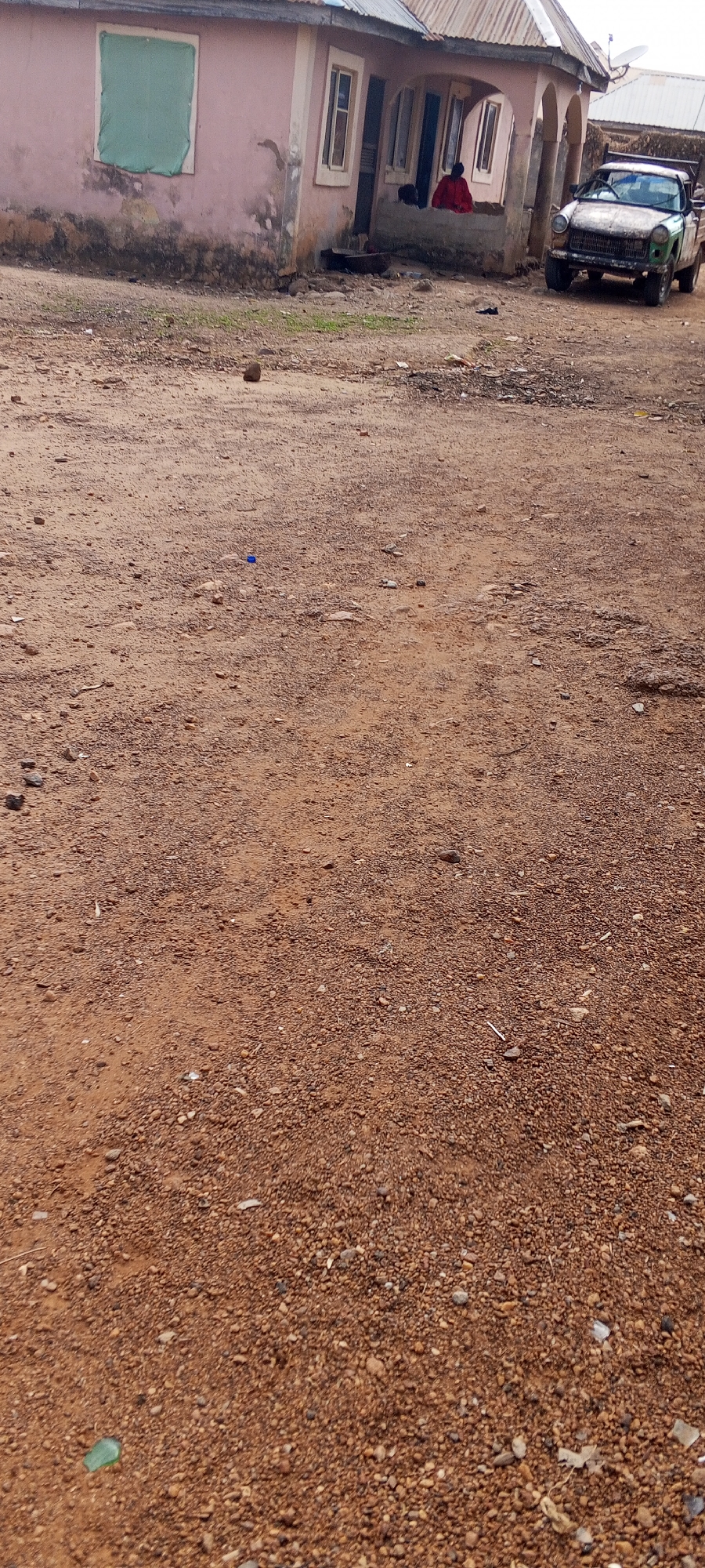 The scene depicts a dirt yard in front of a single-story, pink-toned building in Keffi, Nigeria. A person in a red garment sits on the porch. An old, green pick-up truck is parked to the right of the building. The yard is covered in reddish-brown dirt and small stones, with scattered debris. A small rock and a blue bottle cap are visible on the ground. A green sheet covers a window on the building's left side. Satellite dishes are visible on a nearby roof.