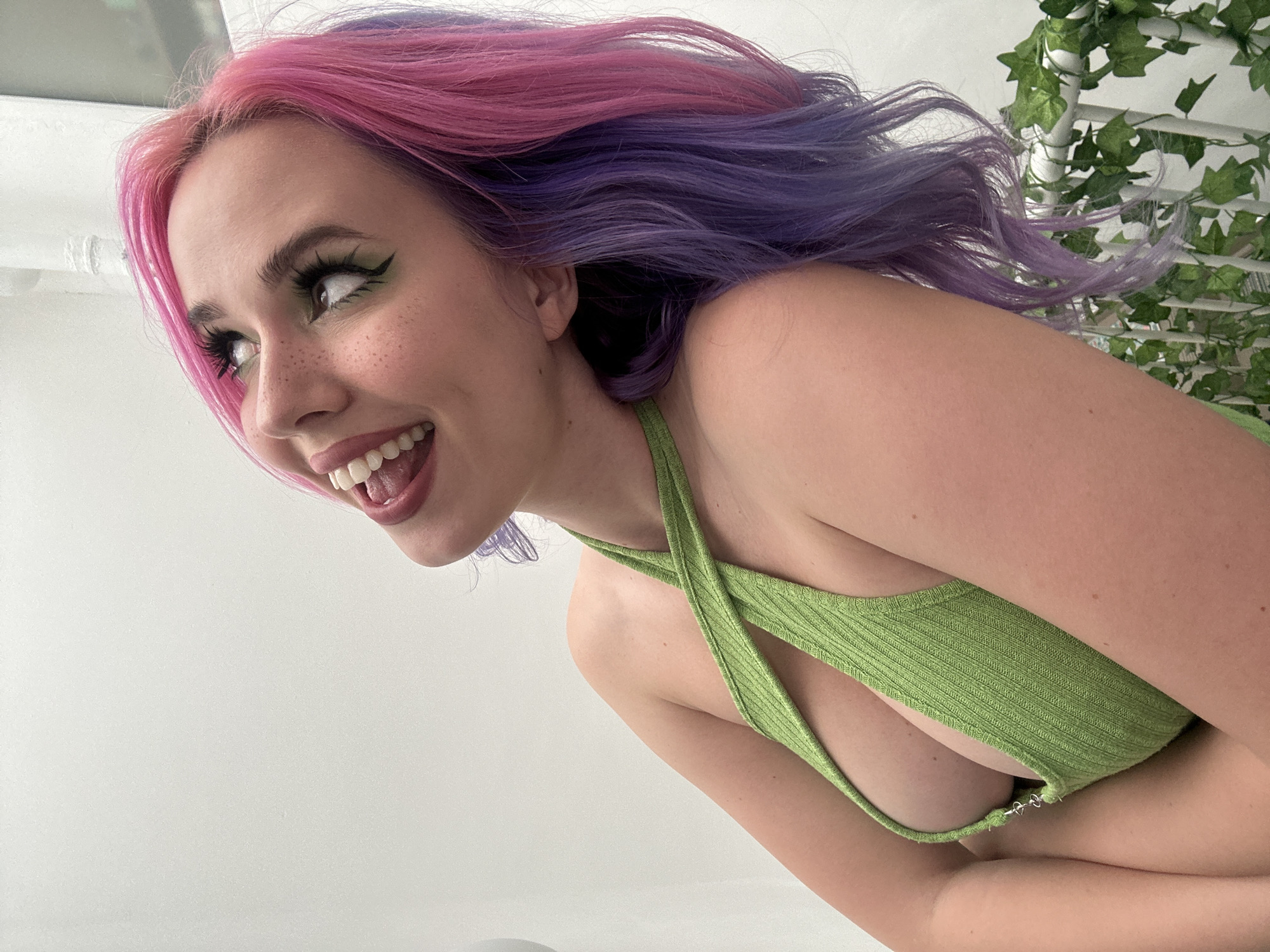 A young woman with pink and purple hair is smiling and looking to her left. She is wearing a light green top with a crisscross design over her chest. In the background, there is a white wall and artificial vines. The location is unknown.
