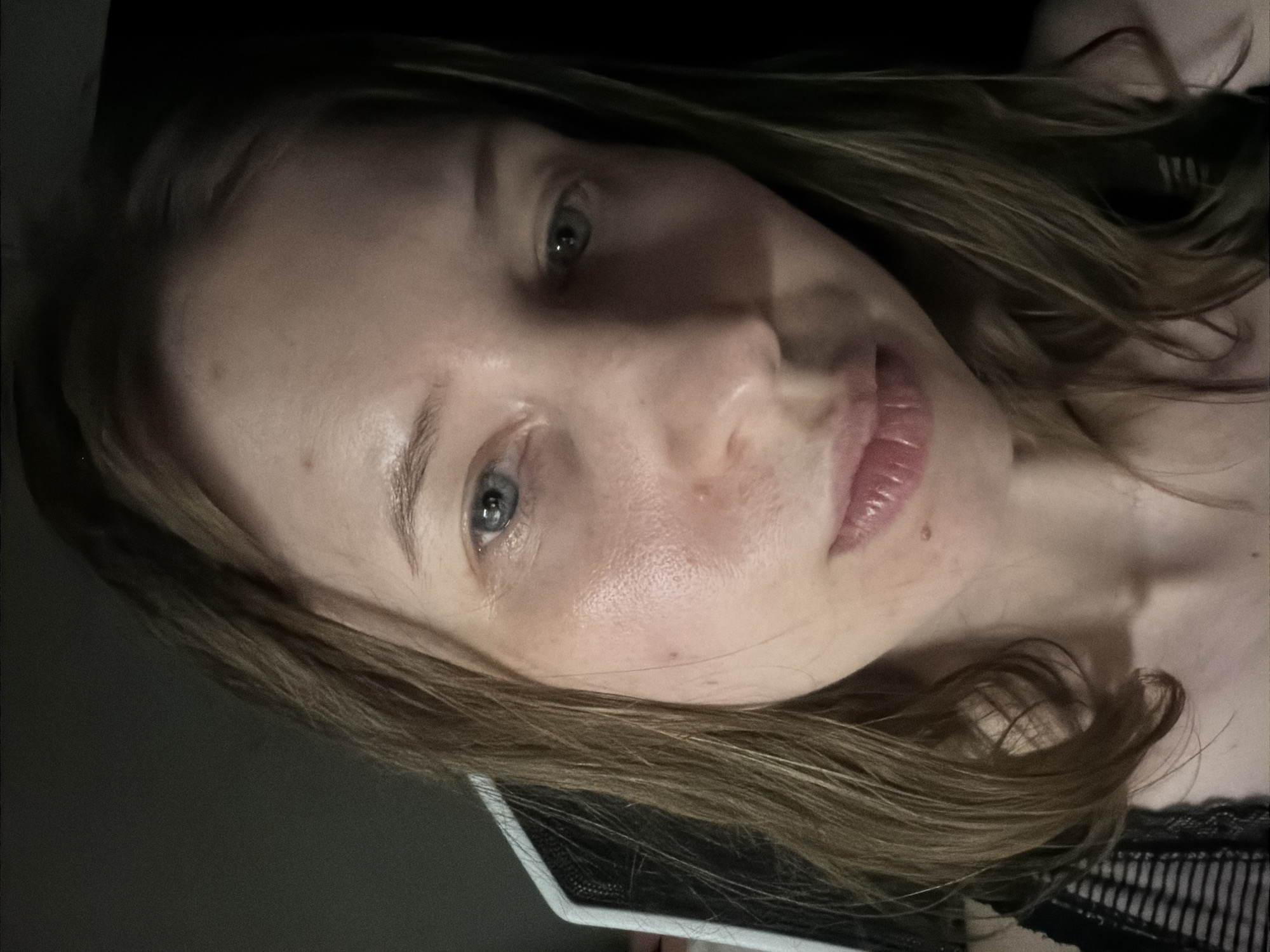 The image shows a close-up of a young Caucasian woman's face. She has blue eyes and light brown hair that is parted in the middle and falls around her shoulders. Her skin appears fair with some redness on her cheeks. She is looking slightly down and to the right. The lighting is dim, with a dark background behind her. To the left of her face, a white object with a black mesh pattern is partially visible, which could be part of a computer monitor or speaker. The woman is wearing a black top with thin straps. The location is not discernible from the image.