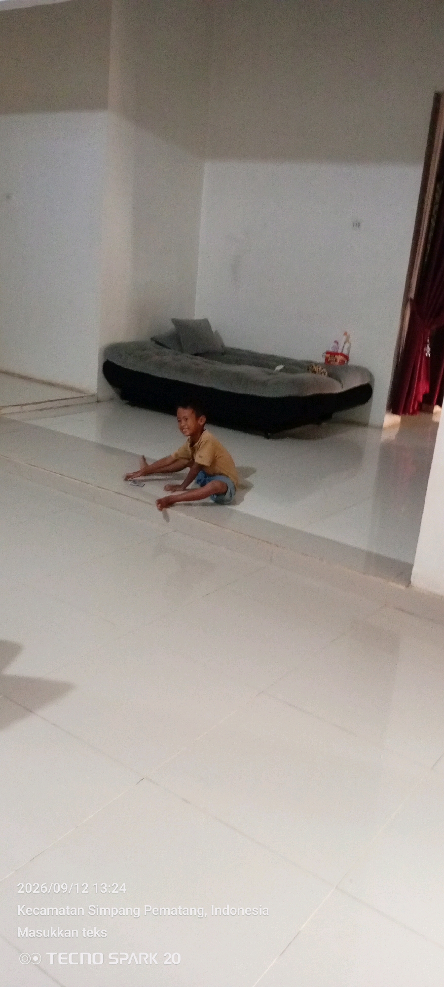 A young boy wearing a yellow t-shirt and blue shorts sits on a light-colored tiled floor in what appears to be a living room. He is facing towards the viewer, with his legs extended in front of him. He is smiling and looking at something on the floor, possibly cards, as his hands are positioned near them. Behind him, an inflatable mattress with two pillows is placed against a white wall. The room has white walls and a continuation of the light-colored tile flooring. A dark red curtain is visible on the right side of the image. The timestamp indicates the image was taken on September 12, 2026, at 13:24, in Simpang Pematang, Indonesia.