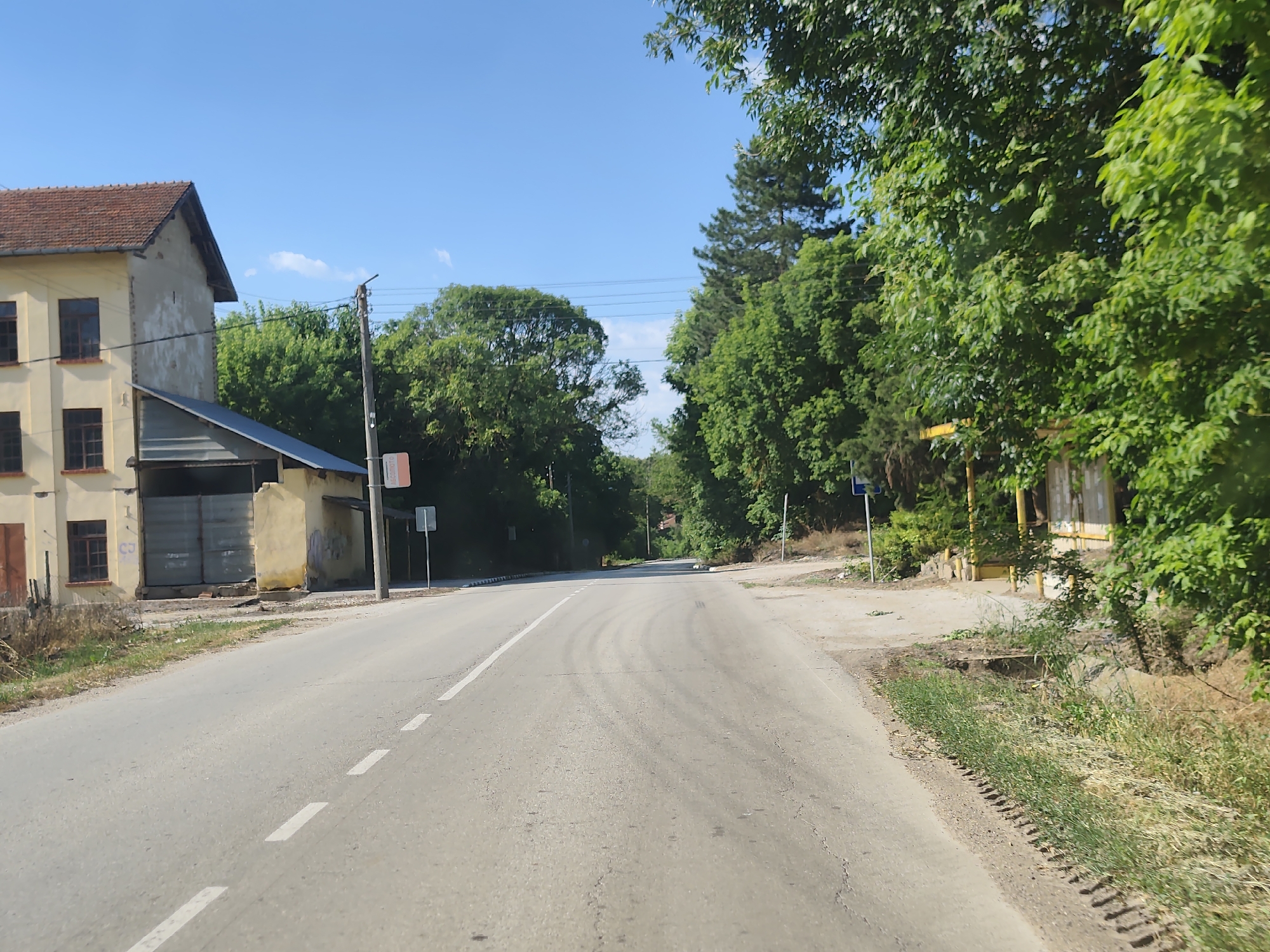 The image depicts a road in Strezherovo, Bulgaria, on a clear, sunny day. A two-lane asphalt road stretches into the distance, bordered by green trees and vegetation on both sides. On the left, a two-story building with a tiled roof and yellow walls is visible, featuring multiple windows and a large, corrugated metal garage door. A smaller, attached structure with a dark roof is also present. Several utility poles and wires are strung along the road. On the right side of the road, a yellow bus stop shelter or similar structure with a wooden frame and a yellow roof is partially obscured by dense green foliage. The roadside is lined with grass and some dry vegetation. The sky is bright blue with a few wispy white clouds. No people are visible in the image.