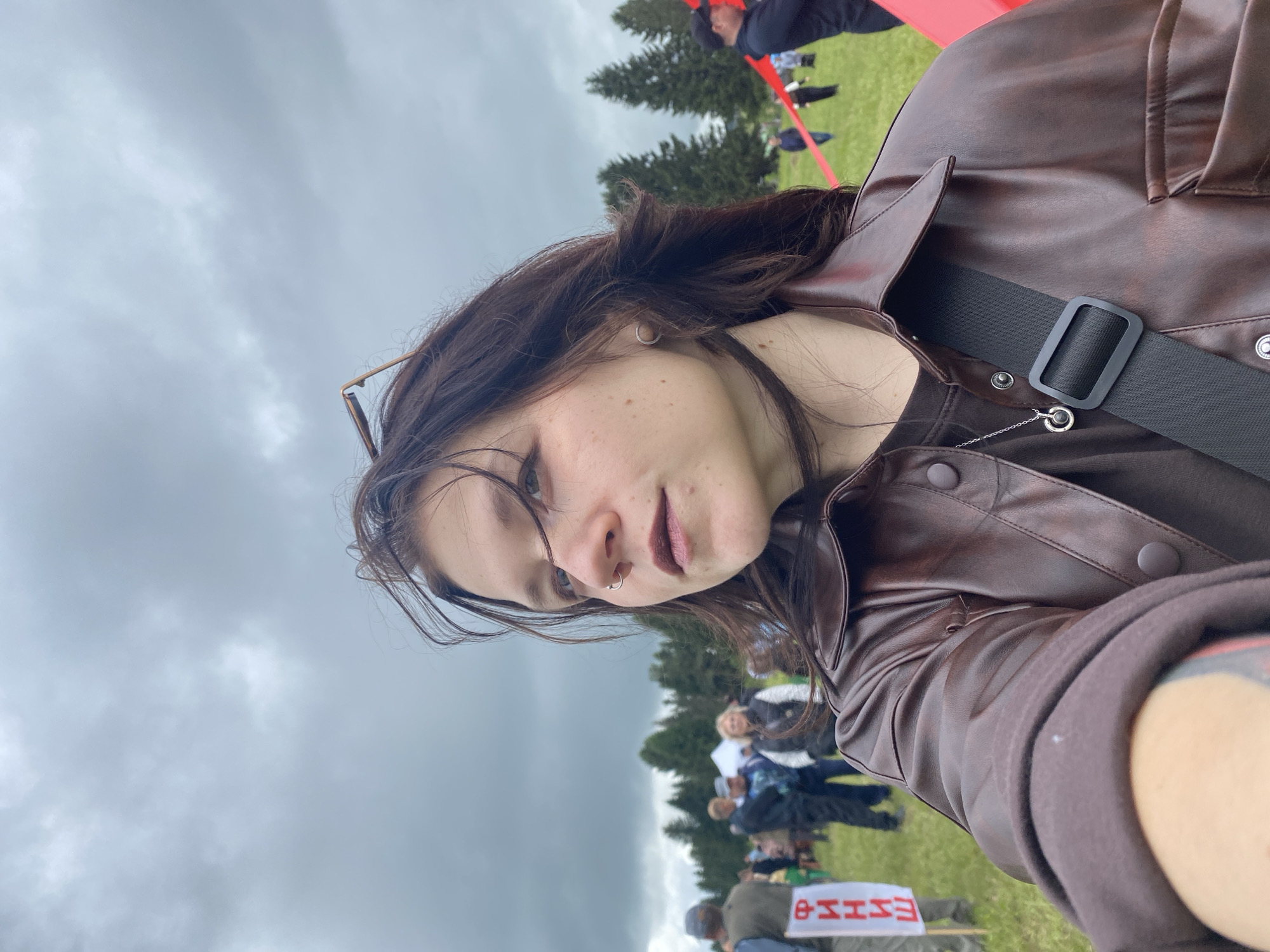 A selfie-style photograph depicts a young adult, likely female, in the foreground, centered slightly to the right, in an outdoor setting in Фоки, Russia. The subject has dark, shoulder-length hair, a nose ring in her left nostril, and dark lipstick. She wears a brown, collared, button-down jacket over a dark t-shirt, a silver necklace with a dark circular pendant, and a black cross-body strap. Sunglasses are perched on her head. Her left arm, partially visible in the lower left, shows a tattoo.
The background reveals a large, open grassy field with scattered individuals and groups. On the far left, a white banner with the red Cyrillic word "ФИНИШ" (FINISH) is visible. In the mid-ground, multiple people are present, some near a white tent. Tall coniferous trees line the distant horizon. A red ribbon or tape extends across the mid-ground on the right side of the frame. The sky is heavily overcast with dense, grey clouds, indicating diffused light and potentially inclement weather.