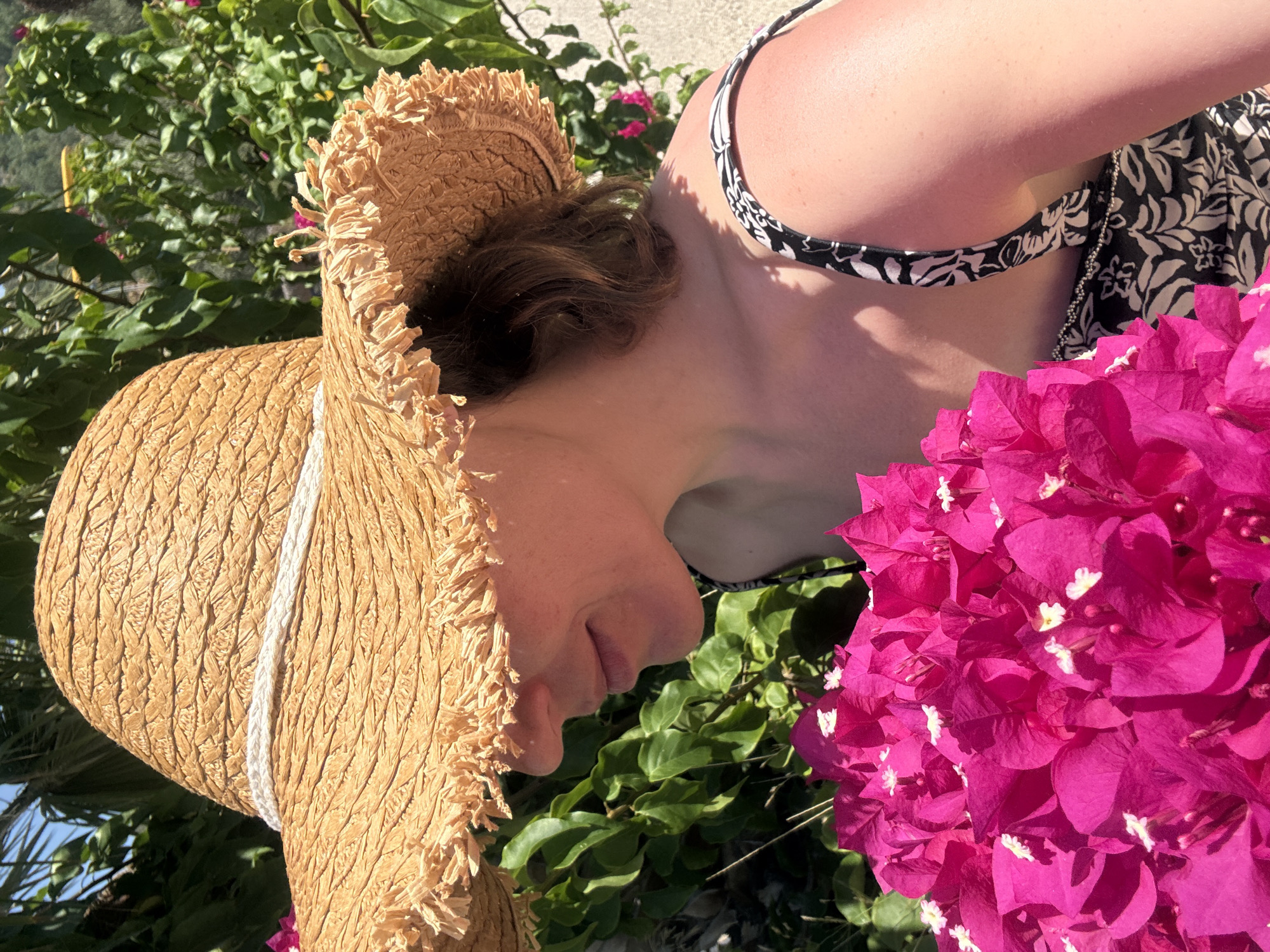 A woman wearing a straw hat and a black and white patterned top is outdoors, holding a large bunch of bright pink flowers. She is positioned in the foreground, with lush green foliage and a hint of blue sky in the background. The image appears to have been taken in Bodrum, Turkey, suggesting a warm, sunny environment suitable for outdoor activities and floral displays common in the region's gardens and bougainvillea-covered walls.