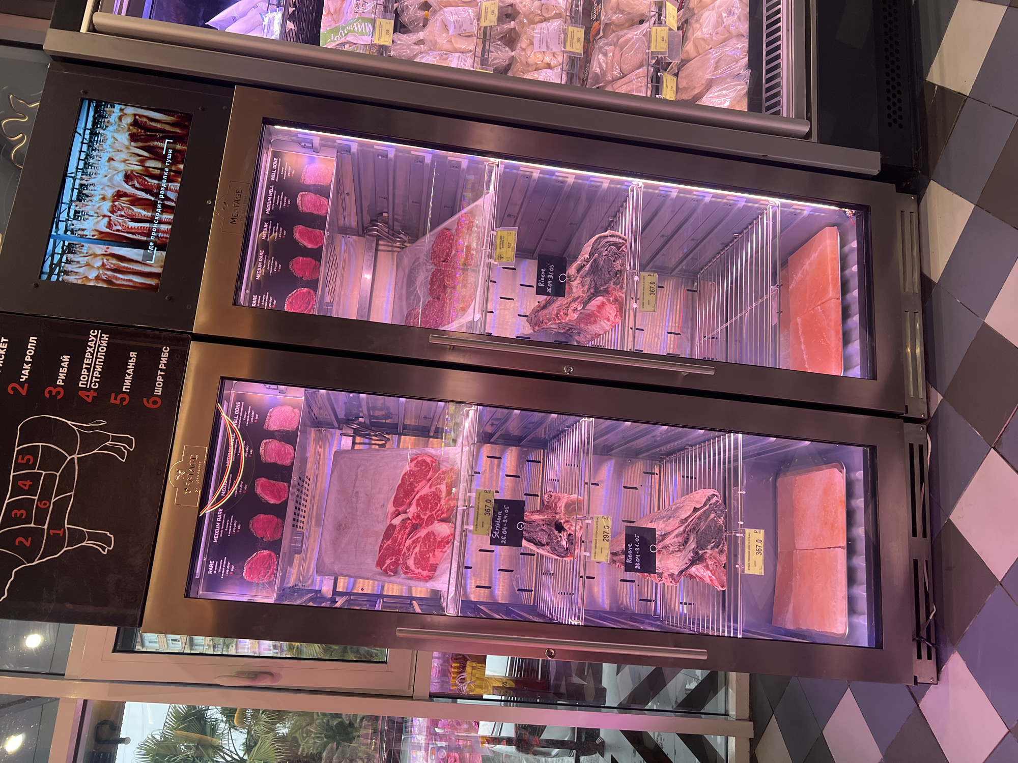 The image shows a refrigerated display case in what appears to be a high-end butcher shop or grocery store, likely in Sochi, Russia, based on the Cyrillic text on the product labels and diagrams. The case contains various cuts of raw meat, primarily beef, including ribeye, striploin, and other cuts as indicated by the meat diagram. Price tags are visible near the meats, and the cuts are neatly arranged in a visually appealing manner. A screen shows how the animal is cut. A sign of rare, medium rare, medium, medium well, and well done is also shown. Above the display case, there are more meats for sale. The floor is tiled with black and white squares. Outside the window is a building and palm trees.