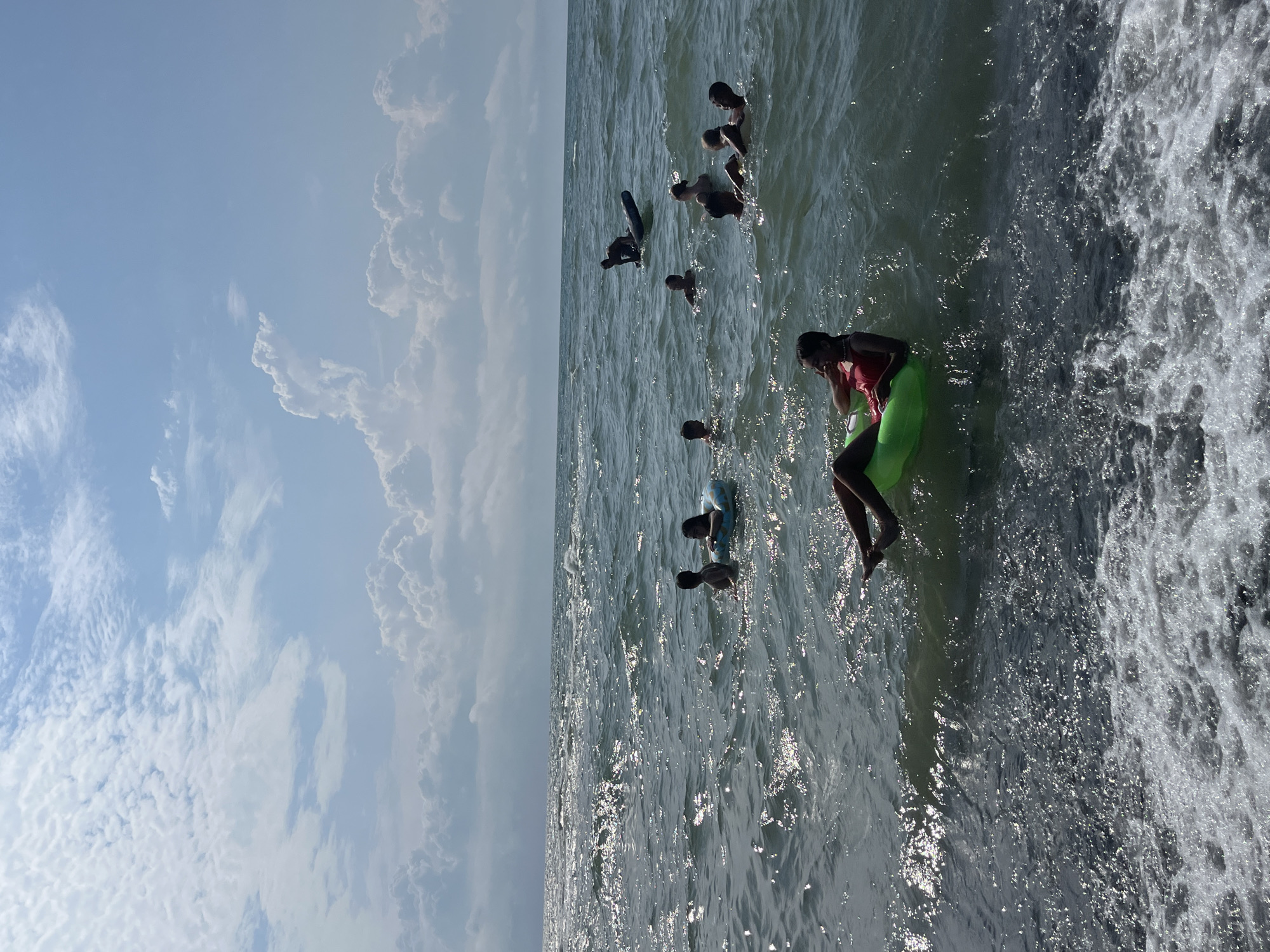 The image captures a sunny day at Ureki, Georgia, where several people are enjoying the sea. The water is a deep blue-green, with moderate waves reflecting the sunlight. In the foreground, a young person is relaxing on a bright green inflatable tube, wearing a red top. Further out in the water, a group of individuals are swimming and playing. One person is on a dark surfboard, while others are in groups, some using inflatable rings. The sky above is a clear blue, dotted with fluffy white clouds, indicating pleasant weather. The overall scene depicts a recreational day at the beach.