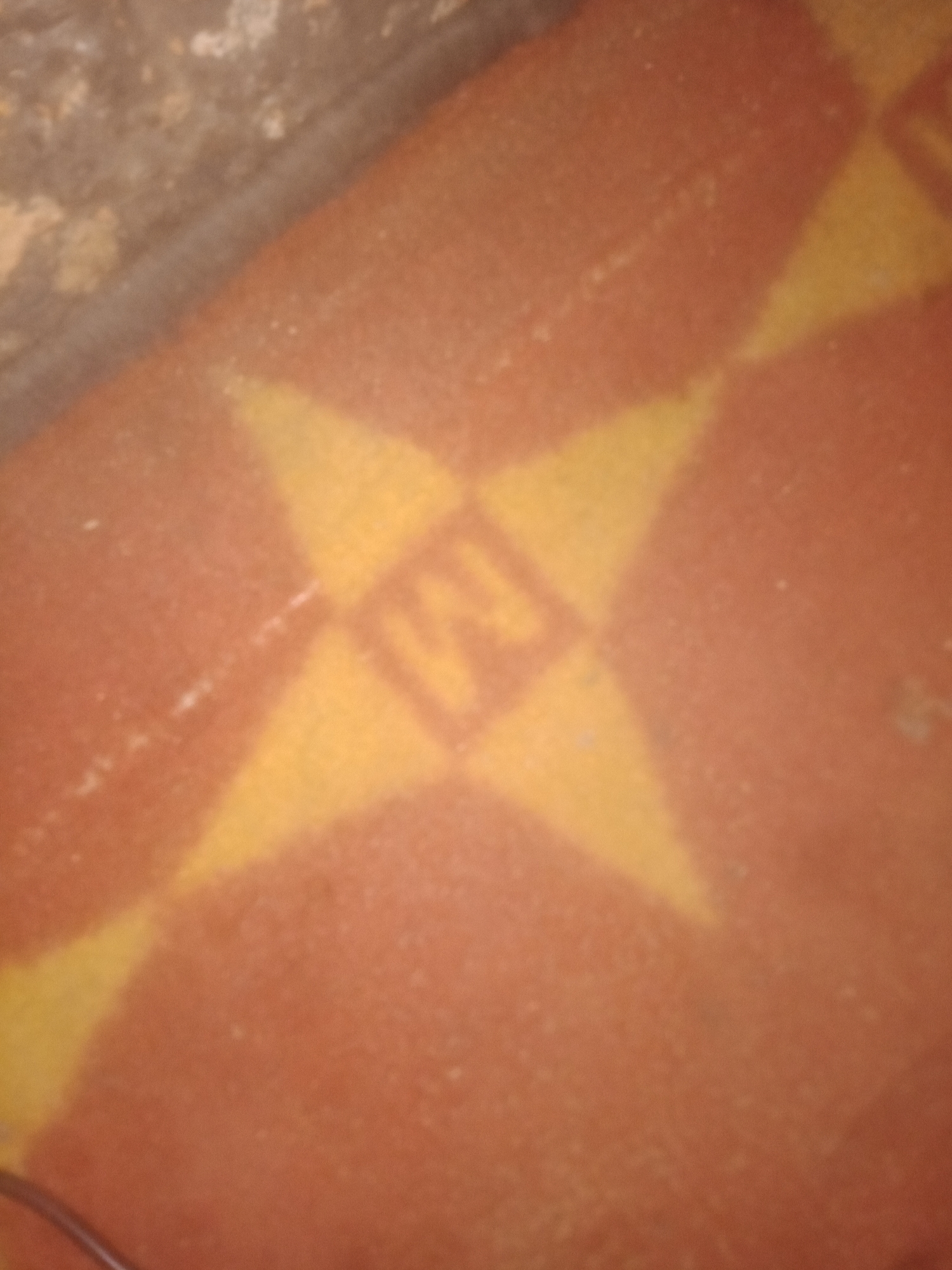 The image shows a close-up, out-of-focus view of a patterned surface, possibly a rug or mat. The pattern consists of repeating diamond shapes in yellow against a reddish-orange background. In the center of one of the yellow diamonds, there appears to be a faint, stylized letter "M". The surface is slightly textured, and there are some small white specks visible. The top left portion of the image is darker and appears to show a rougher, possibly stone or concrete texture, meeting the patterned surface at an angle. A thin black wire or cord is partially visible in the bottom left corner. The lighting is dim, and the image quality is poor, with significant blur and noise. No people or specific urban environment details are discernible.