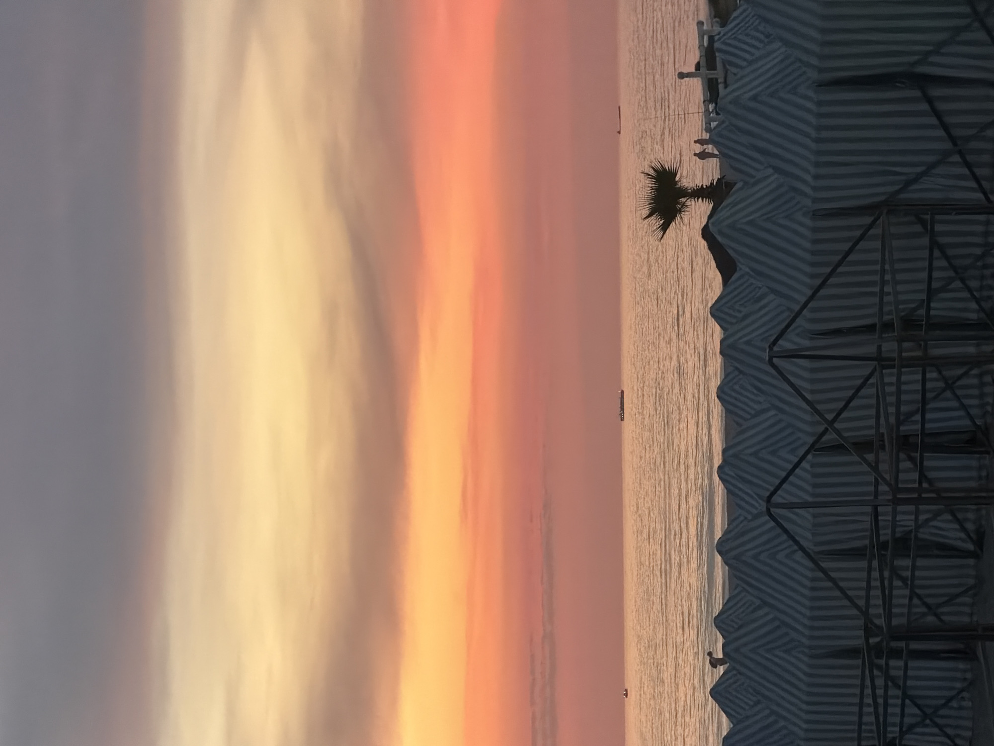 The image depicts a serene sunset scene at Praia da Baía in Espinho, Portugal. The sky is a gradient of colors, transitioning from a soft grey at the top to vibrant oranges and pinks near the horizon. The calm sea reflects these colors, with a few ships visible in the distance. In the foreground, a row of striped tents covers the sandy beach, with a single palm tree standing tall near the waterline. A few people are scattered along the beach, enjoying the view. The overall atmosphere is peaceful and tranquil.