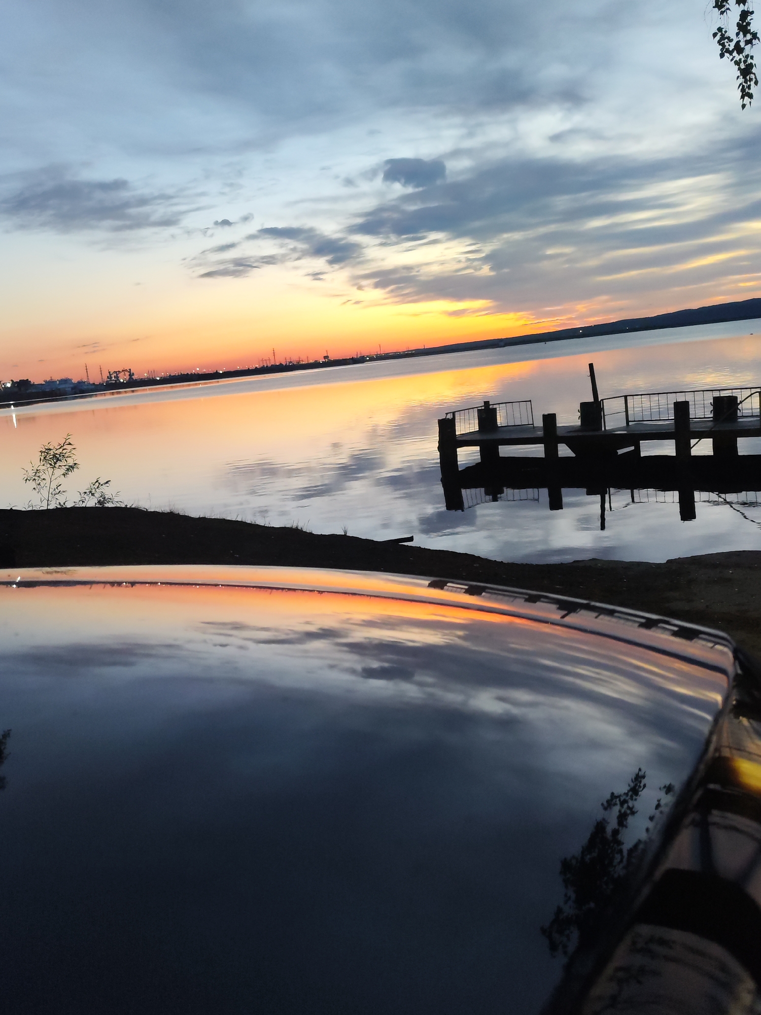The image depicts a sunset over the Kama River in Chaikovsky, Russia. The sky is a mix of orange, yellow, and gray clouds, reflected in the calm water. In the middle ground, there is a small pier with dark wood structures. The shoreline is visible with some vegetation. In the foreground, there is a dark, reflective surface of what appears to be the roof of a vehicle, also mirroring the sunset and sky. There are no people visible in the image. The industrial skyline is visible in the distance.