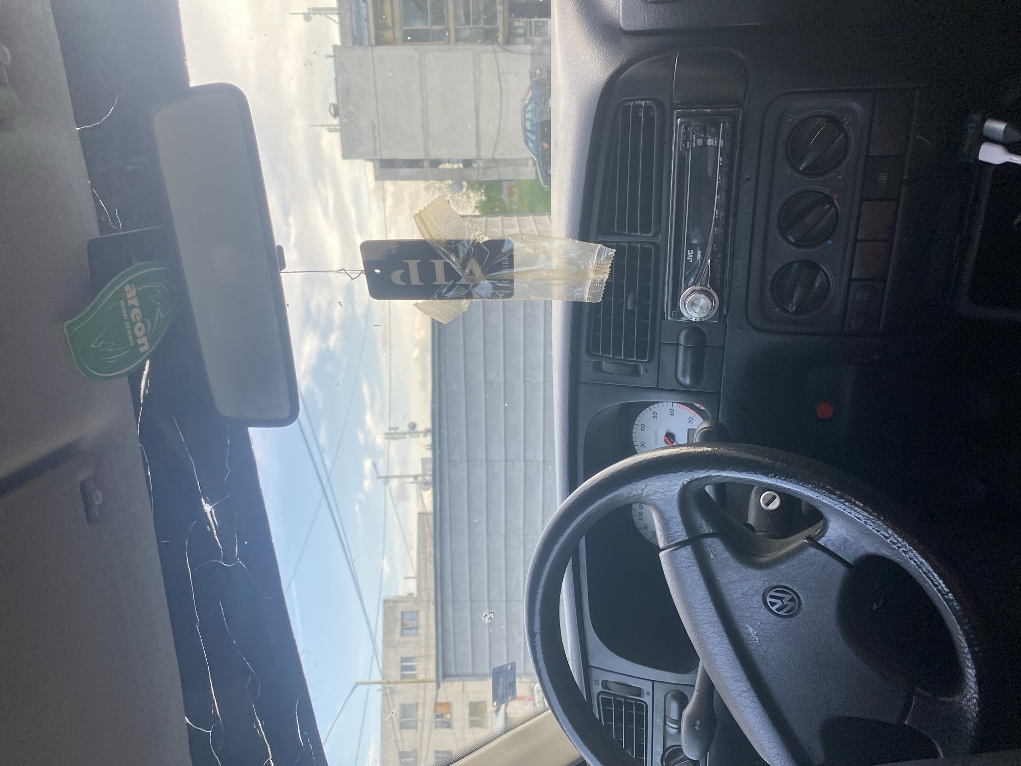 The image is taken from inside a car, focusing on the dashboard, steering wheel, and the view through the cracked windshield. A VIP tag hangs from the rearview mirror, along with an "Areon" air freshener. The car's dashboard features a JVC radio and ventilation controls. The steering wheel has a Volkswagen logo. Through the windshield, buildings and a street can be seen, suggesting an urban setting. The location is identified as Debelt, Bulgaria, based on the VIP tag which is a product of the company Gesiba there. The windshield damage obscures some of the view.