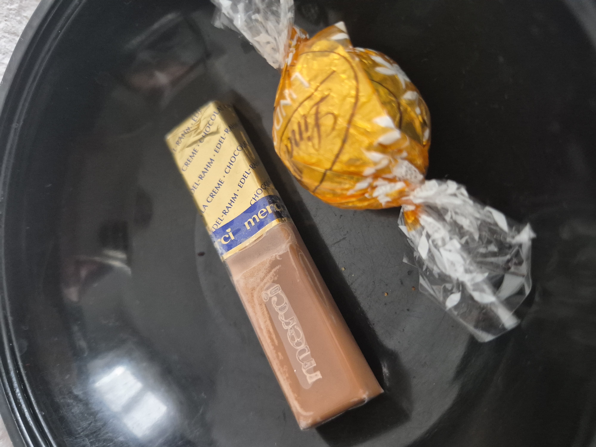 An overhead, slightly angled medium shot captures two chocolates resting in a dark, circular bowl. To the left, a single, elongated chocolate bar, wrapped in clear plastic, lies horizontally. The word "merci" is embossed into the surface of the chocolate. To the right of the bar, a spherical chocolate, wrapped in crinkled golden foil with white floral patterns, is positioned at an angle, its plastic wrapper extending towards the bottom left. The bowl's surface is dark and slightly reflective, showing some smudges and scratches.