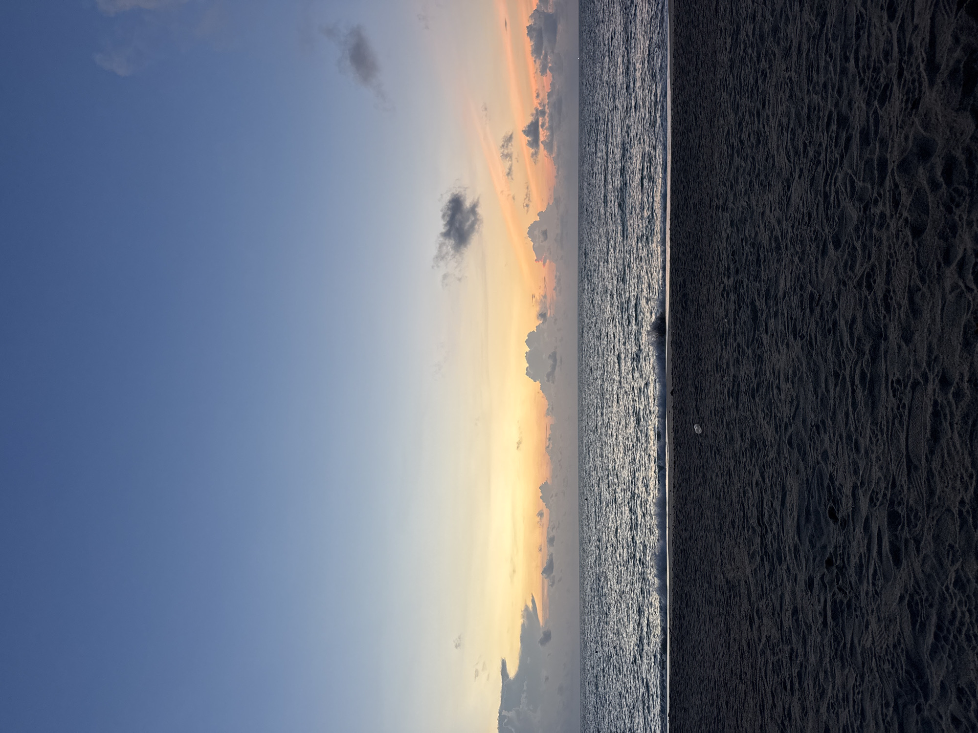 The image shows a beach scene in Baan Thong Nang, Thailand, taken at sunrise or sunset. The sky transitions from blue to orange and yellow hues near the horizon, with scattered clouds. The ocean occupies the central portion of the frame with visible wave patterns. The right side of the image is dominated by dark sand with textured patterns. There are no people visible in the shot.