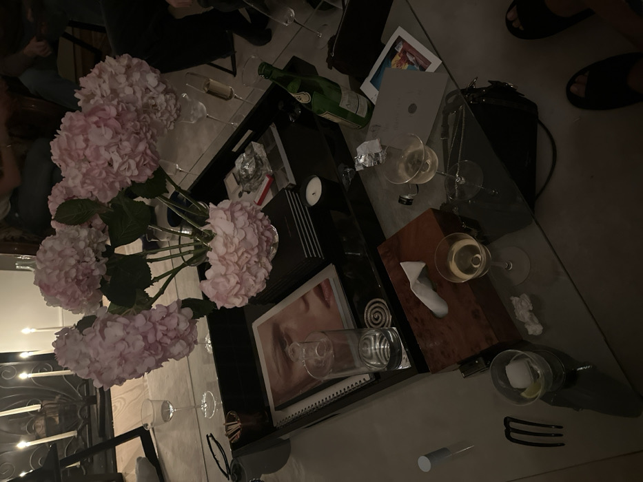 The scene depicts an indoor, low-lit domestic setting, located in the city of London, United Kingdom, likely an evening social gathering. The primary focus is a glass coffee table laden with various items.
At the center of the table, a large, dark rectangular tray holds a clear glass vase containing multiple large pink hydrangea blossoms. On the tray, an open magazine displays a close-up image of a person's face, resting beside a stack of dark-bound books. A clear glass ashtray with a cigarette butt and ash, a red matchbox, a small unlit candle, and a tall glass of water on a spiral-patterned coaster are also present on the tray.
Surrounding the tray on the glass table are additional objects: a dark wooden tissue box with a single white tissue protruding, multiple stemmed wine glasses containing varying amounts of white wine or champagne, a short cocktail glass with ice and a lime wedge, and a green glass bottle (partially consumed, possibly Perrier). A small black handbag or clutch, a black three-pronged hair comb, reading glasses, and crumpled white paper are also visible.
In the background, out of focus, multiple individuals are partially visible, seated, suggesting a group of people. Two feet, clad in dark open-toed sandals, are visible in the bottom right corner, resting near the table. The overall lighting is dim, augmented by multiple lit white tapered candles in a candlestick holder visible in the upper left corner, situated in front of a dark, possibly metal, fireplace screen.