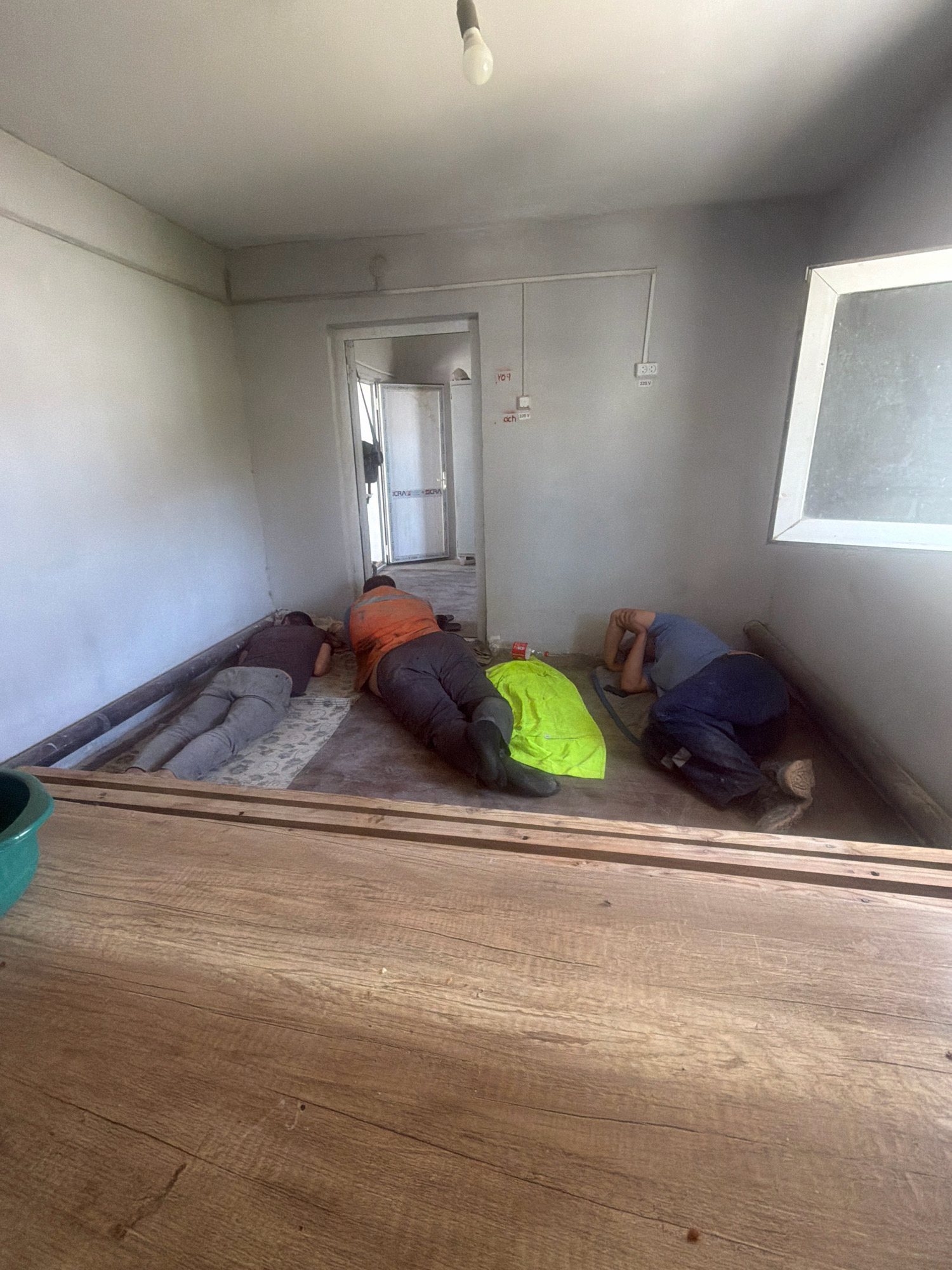 Four individuals are lying on the floor in what appears to be an unfinished room. The room has gray walls, a white ceiling with a visible light fixture, and a doorway leading to another area. The floor is partially covered with a patterned rug and has some debris. In the foreground, there is a wooden surface.
The individuals are dressed in work-related attire. One person on the far left is lying on their side, wearing gray pants and a dark shirt. Next to them, a person in gray pants and a dark shirt is lying face down. Further in the center, a person wearing an orange vest over dark clothing is lying face down. On the far right, a person in blue work pants and a blue shirt is lying on their side with their arms over their head. A bright yellow blanket or tarp is positioned between the center and right individuals.
The room appears to be in a state of construction or renovation. Electrical conduits and outlets are visible on the walls. A green bucket is present in the lower left corner of the image. The overall scene suggests a break or rest period for the individuals. There is no indication of the specific location within the city of Besapan, Uzbekistan.
