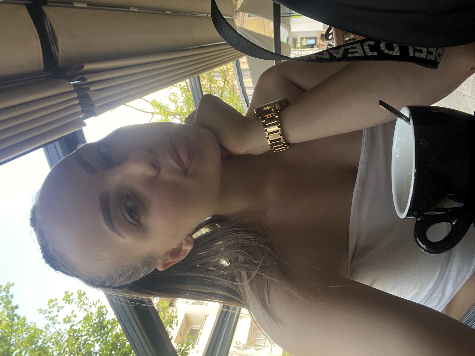 A selfie taken in the city of Troyan, Bulgaria, shows a young woman with light skin, brown hair, and prominent eyebrows. She wears a gold wristwatch on her left wrist and is leaning her chin on her hand. She's wearing a white top. A black mug with a spoon inside sits to her right. A black strap with "DECI JEANS" printed on it is visible near her shoulder. The background features a partially visible awning, a window, green trees, and a building.
