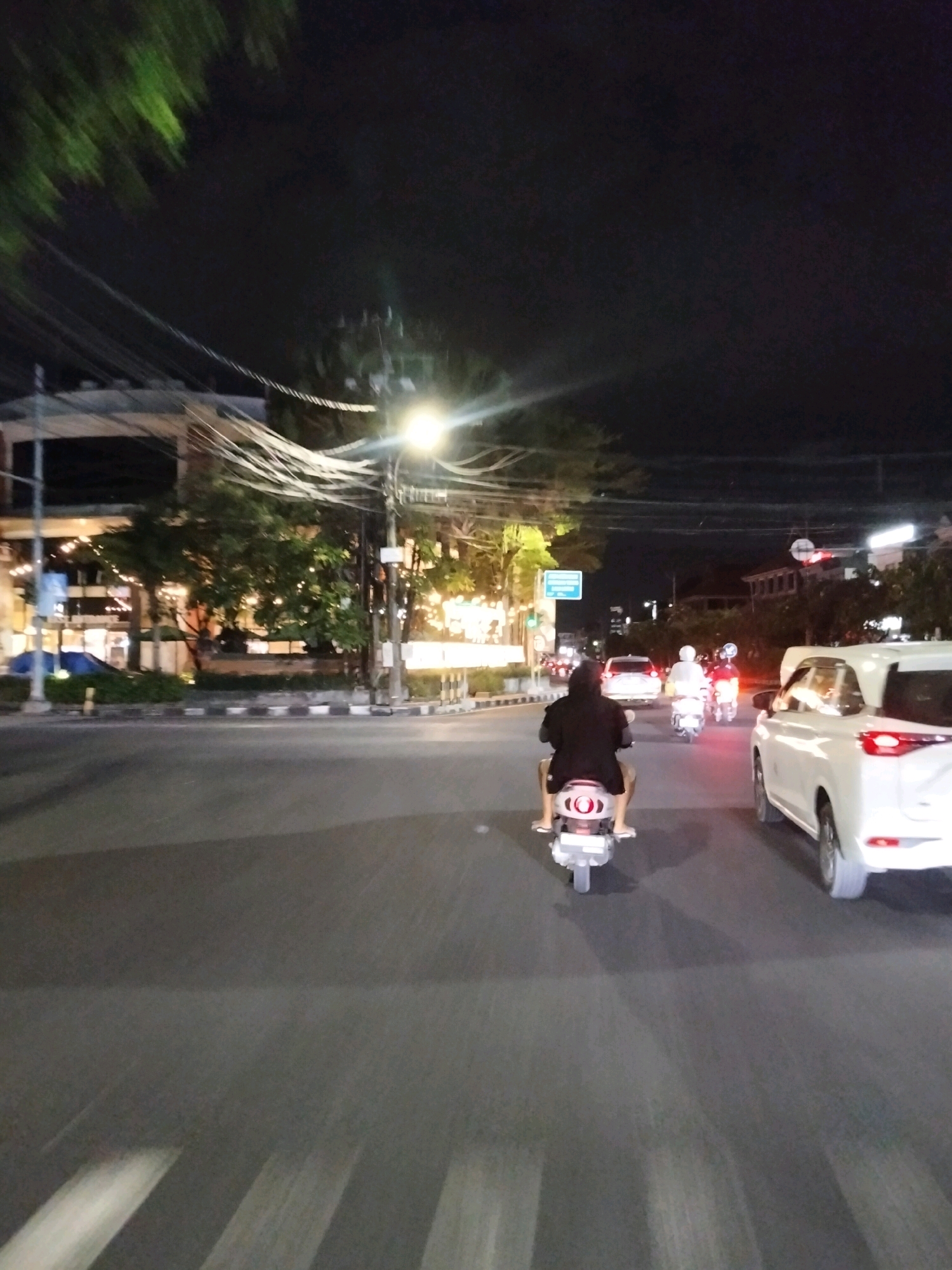 This image captures a street scene at night in Denpasar, Indonesia. In the foreground, a person on a white scooter travels on a paved road, with the scooter's taillight illuminated. To the right, a white SUV is parked or moving slowly, its rear lights visible. Further down the road, other scooters are visible, along with a white car and trees lining the street. The background shows buildings with illuminated windows and a street light with a bright glow. The sky is dark, indicating nighttime.