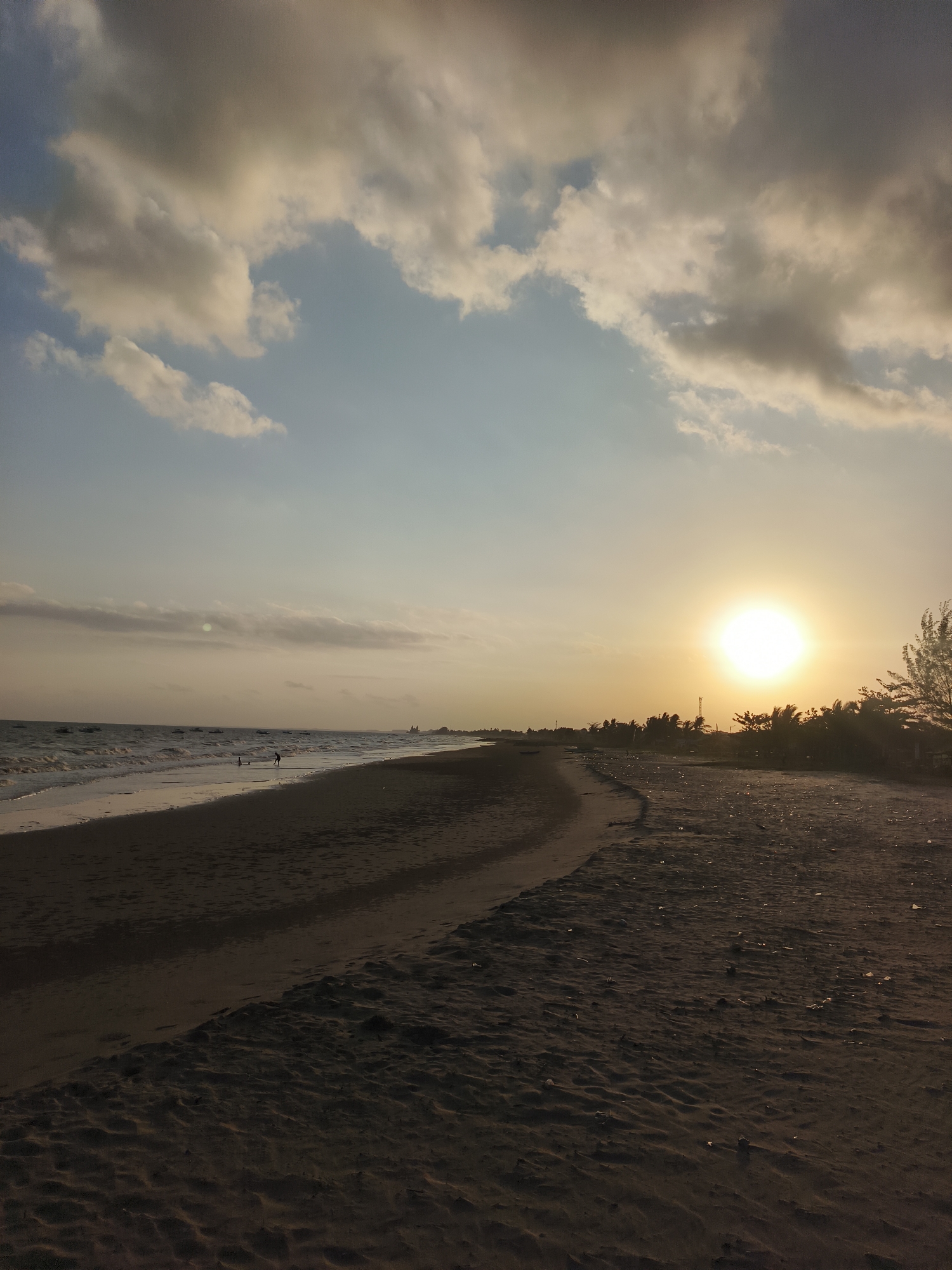 The image captures a serene sunset scene on a beach in Kupang Berkah Jaya, Indonesia. The sky is a gradient of soft blues and oranges, with scattered clouds tinged by the setting sun. The sun, a bright orb, is positioned on the right side of the frame, casting a warm glow across the landscape. A gentle wave line marks the boundary between the calm ocean and the sandy shore. The beach itself is composed of dark sand, with subtle undulations and a faint path winding along its surface. In the distance, the coastline is lined with silhouetted trees and vegetation, with a few boats visible on the water. Two individuals are present on the beach, their figures small against the vast expanse of the natural scenery, appearing to be walking or playing near the water's edge. The overall atmosphere is tranquil and natural.
