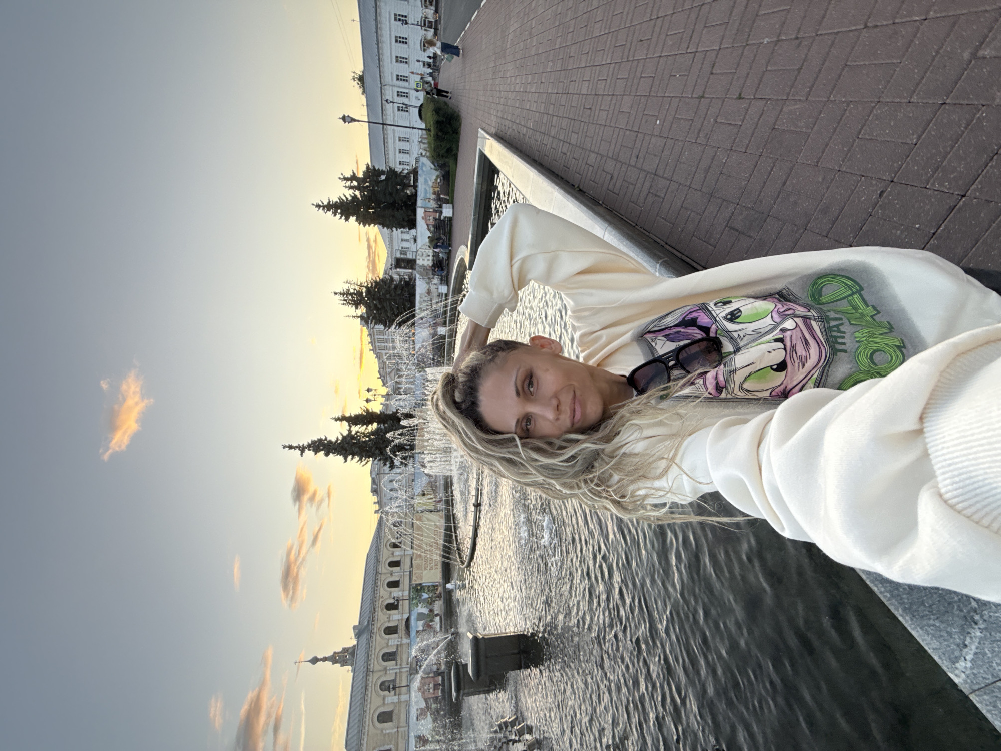 In Rusanovo, Russia, a woman with long, curly blonde hair styled in a high ponytail takes a selfie in front of a large, ornate fountain. The fountain has multiple tiers and spouts water into a large pool. The woman is wearing a light-colored hoodie with a cartoon character graphic and sunglasses perched on her neckline. Behind the fountain, a collection of buildings with classical architecture are visible, some with arched windows and a prominent steeple. Several tall, dark green evergreen trees are scattered throughout the background. The sky is a soft gradient of pale blue and orange, indicating either sunrise or sunset, with a few wispy clouds. The ground in front of the fountain is paved with red brick in a herringbone pattern. To the right of the woman, a paved walkway extends into the distance, with a few pedestrians walking and a lamppost visible. The overall scene is an outdoor urban setting with a historical architectural style, bathed in the soft light of dusk or dawn.