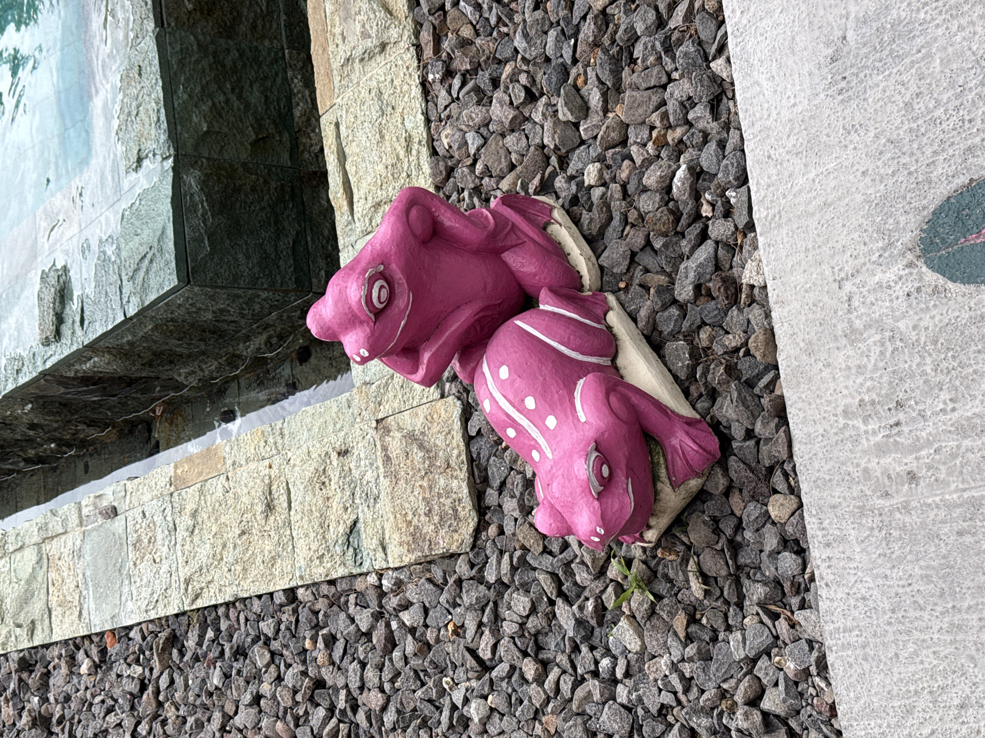 Captured from a high angle, the scene presents a close-up view in what appears to be an outdoor space in Unknown, Indonesia.
Dominating the composition are two pink, frog-shaped statues placed on a bed of small, gray gravel. The statues are positioned next to a structure constructed from light-colored stone, possibly a water feature or part of a garden design. Water is visible in a corner of the structure.
Adjacent to the gravel bed is a light gray slab, potentially a walkway or part of the hardscaping, adorned with a dark green circular pattern. The scene lacks signs of human activity.