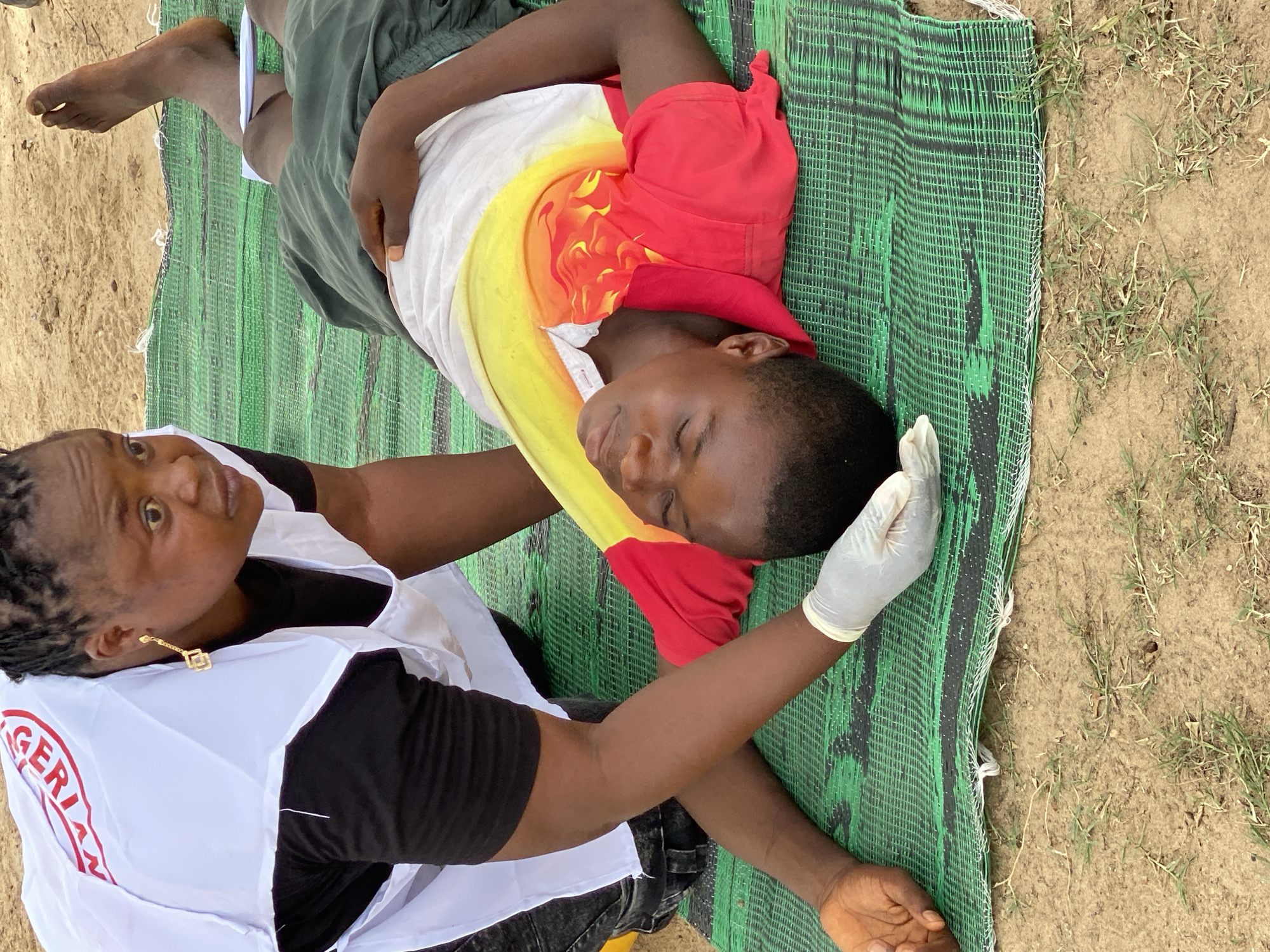 The image shows a person lying on a green mat on the ground, with another person kneeling beside them. The person on the ground is wearing a white t-shirt with a colorful design, green shorts, and is barefoot. The kneeling person is wearing a white vest over a black shirt and has a surgical glove on one hand. The vest has lettering on it that includes the words "NIGERIAN RED CROSS". The surrounding environment appears to be sandy soil with some sparse vegetation. The location is Kinkinau, Nigeria. The person on the ground appears to be receiving medical attention.